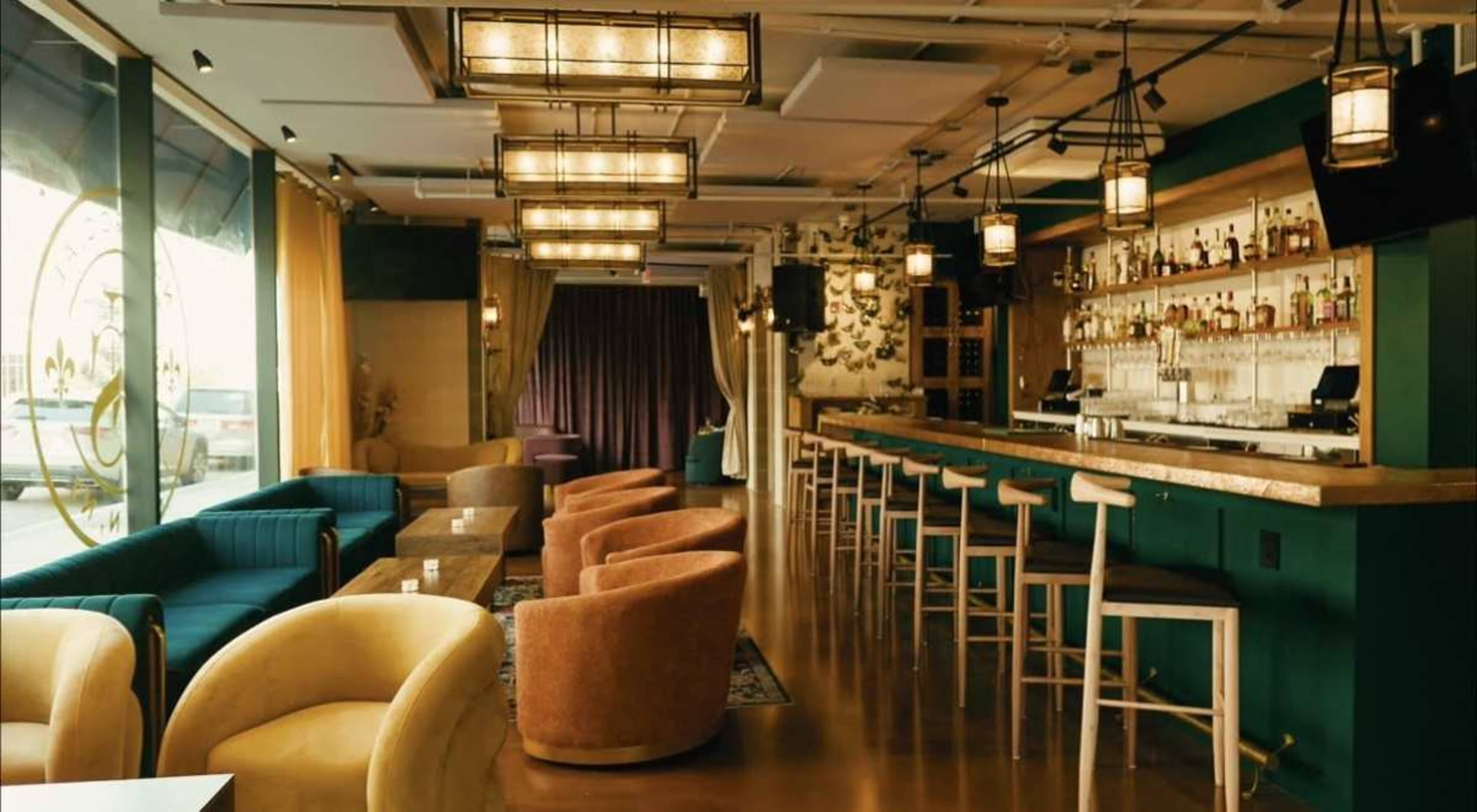 The image shows a modern bar and lounge area with colorful seating options, a well-stocked bar, and large windows allowing natural light to enter.