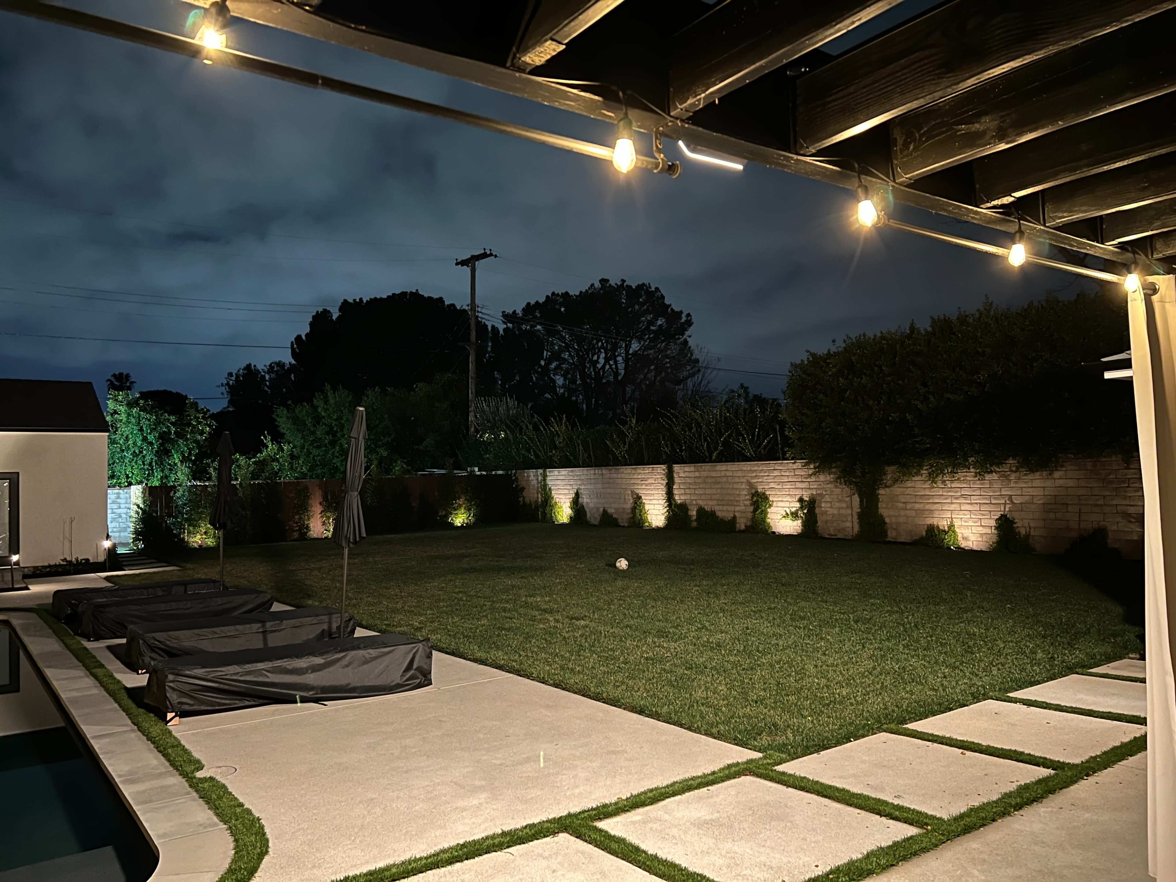 The image shows a backyard at night with a well-maintained lawn, illuminated by string lights and low garden lights along the perimeter.