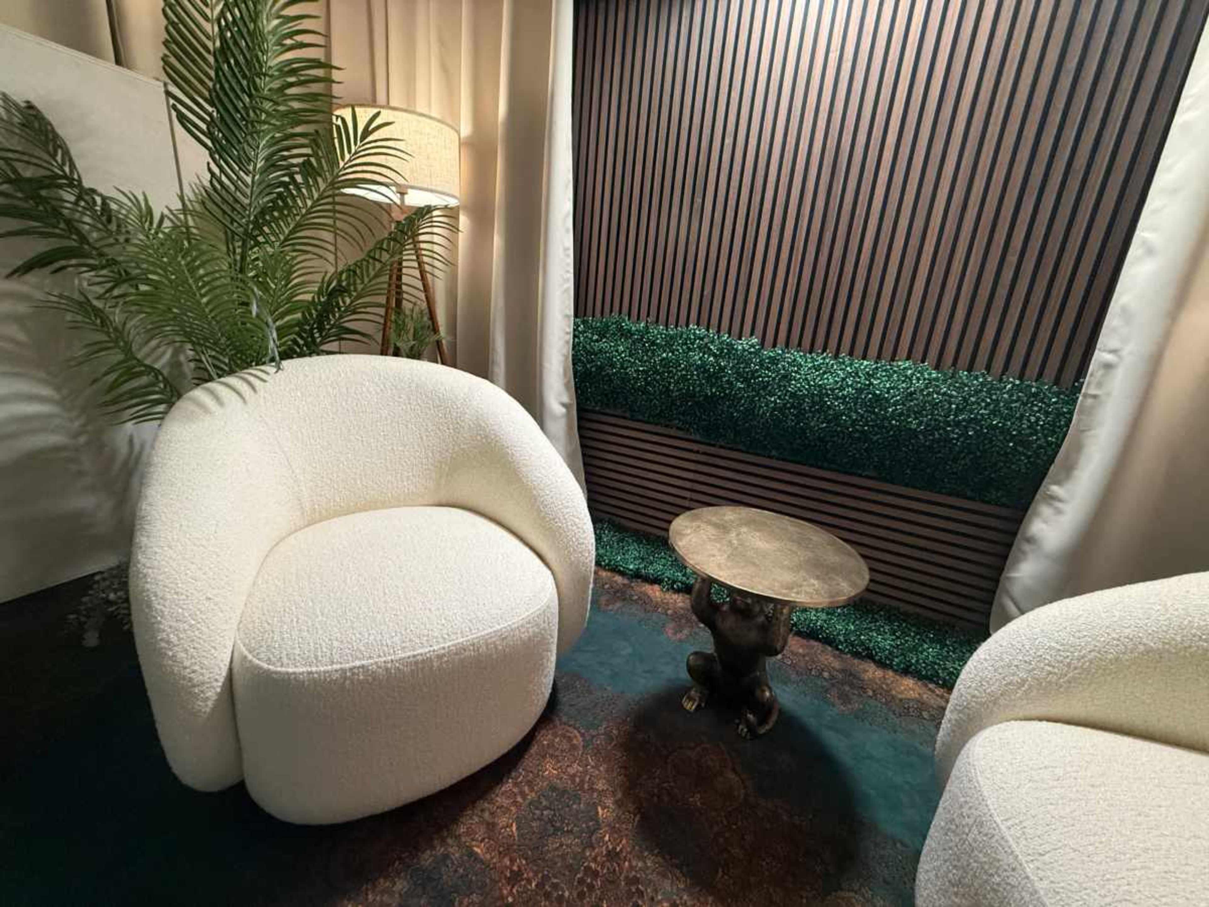 The image shows a cozy seating area with two plush white chairs, a small golden table, and a backdrop of artificial greenery and wooden paneling.
