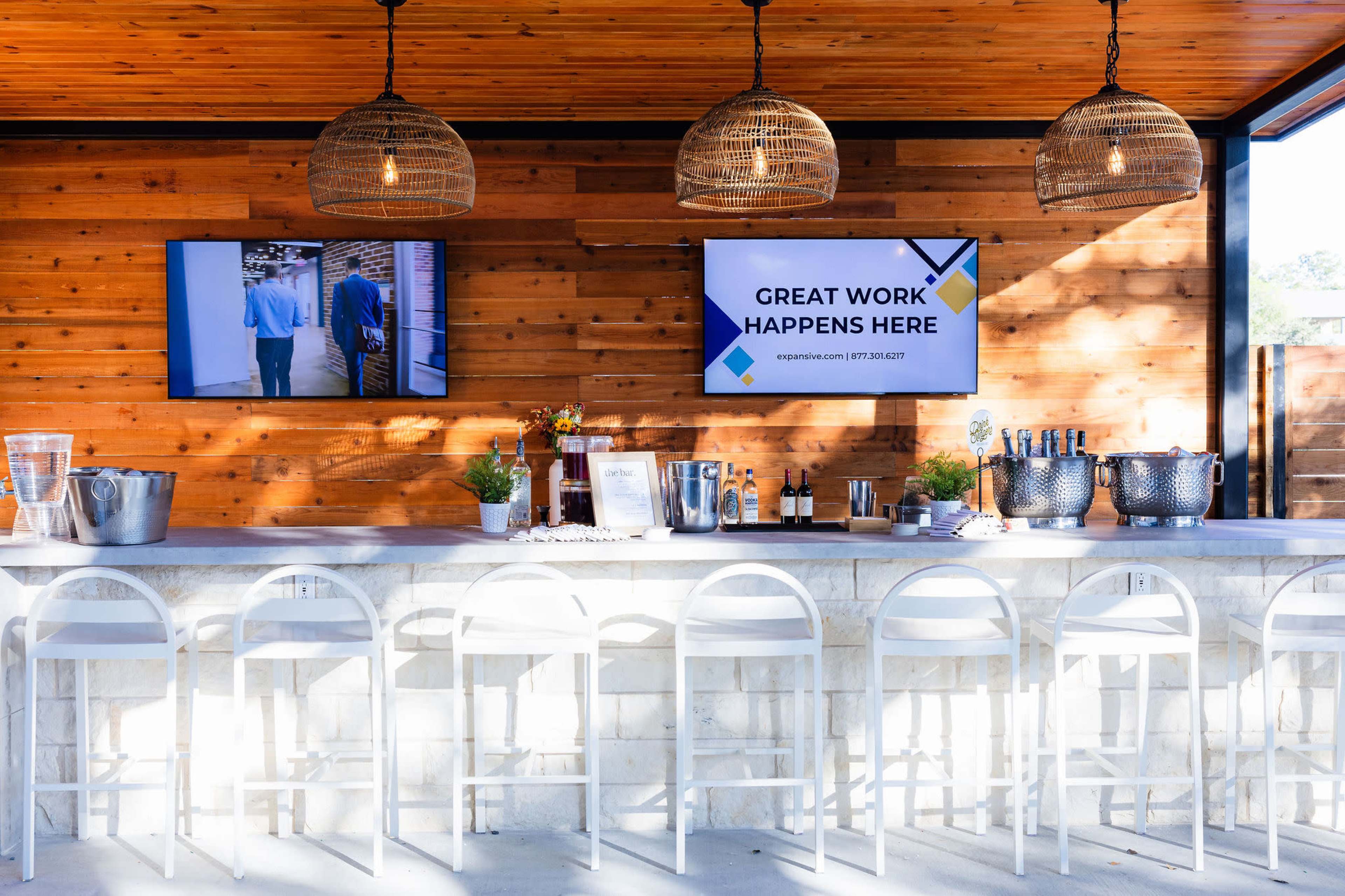 A modern outdoor bar area features a stone counter with white stools, two hanging light fixtures, and two screens displaying promotional content against a wooden wall.