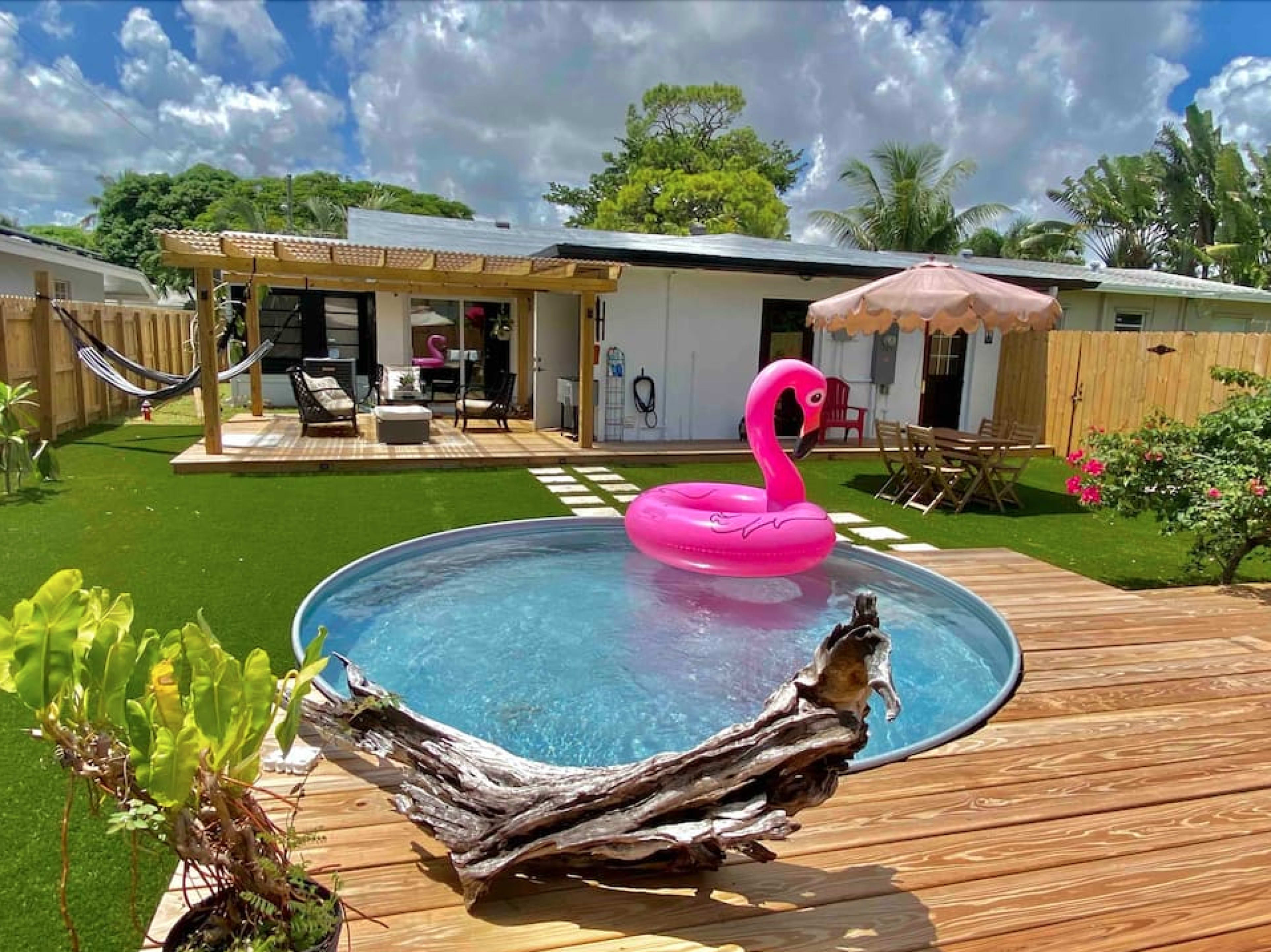 A backyard features a round swimming pool with a pink flamingo float, surrounded by wooden decking, lounge furniture, and a shaded seating area.