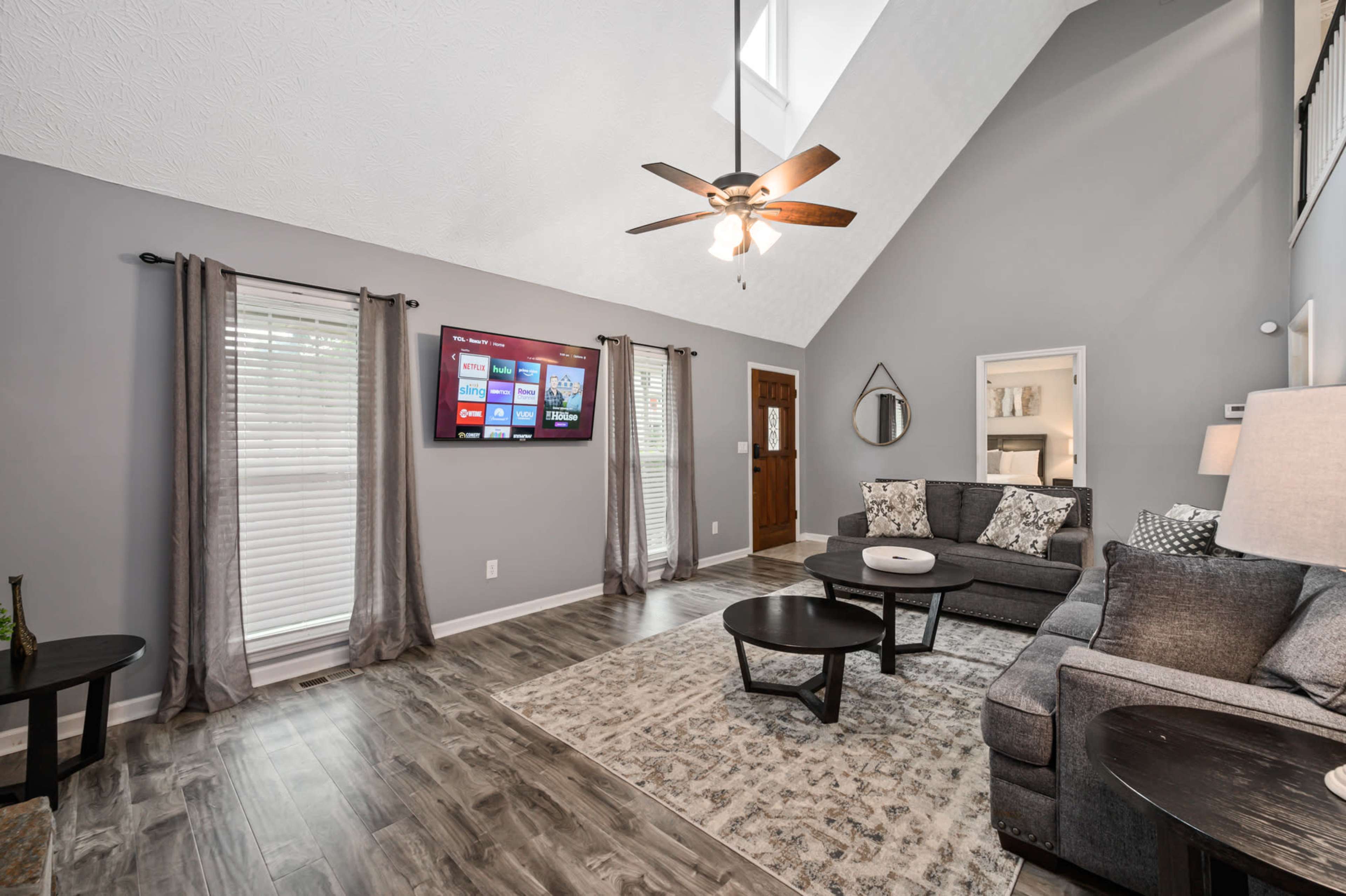 A stylish living room features a gray sectional sofa, a round coffee table, a large wall-mounted TV, and tall windows allowing natural light to enter.