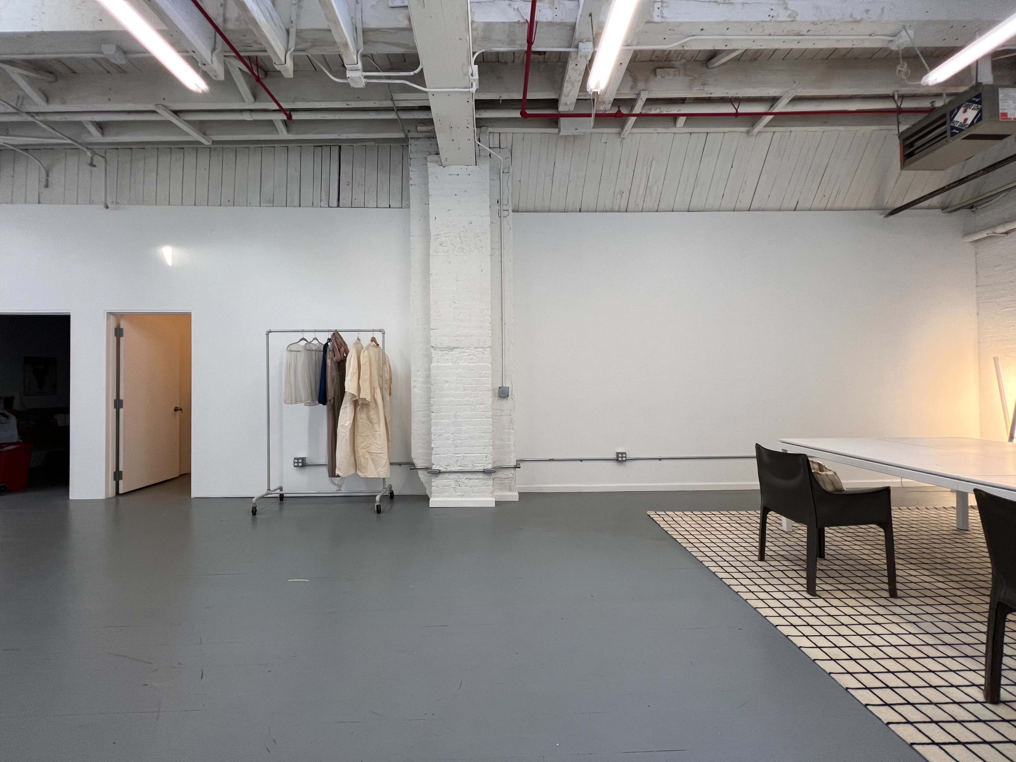 A spacious, minimalistic room with a white wall, a clothing rack with garments, a table, and two black chairs on a tiled rug.