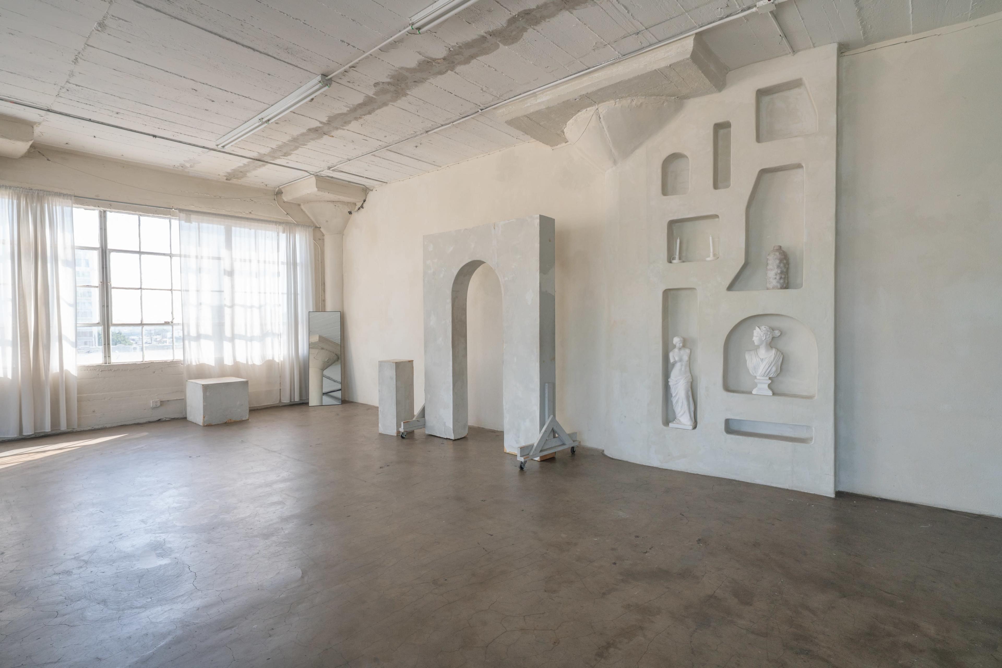 The image shows a spacious, minimalist studio with large windows, showcasing white walls and various architectural features, including an arch and wall niches with sculptures.
