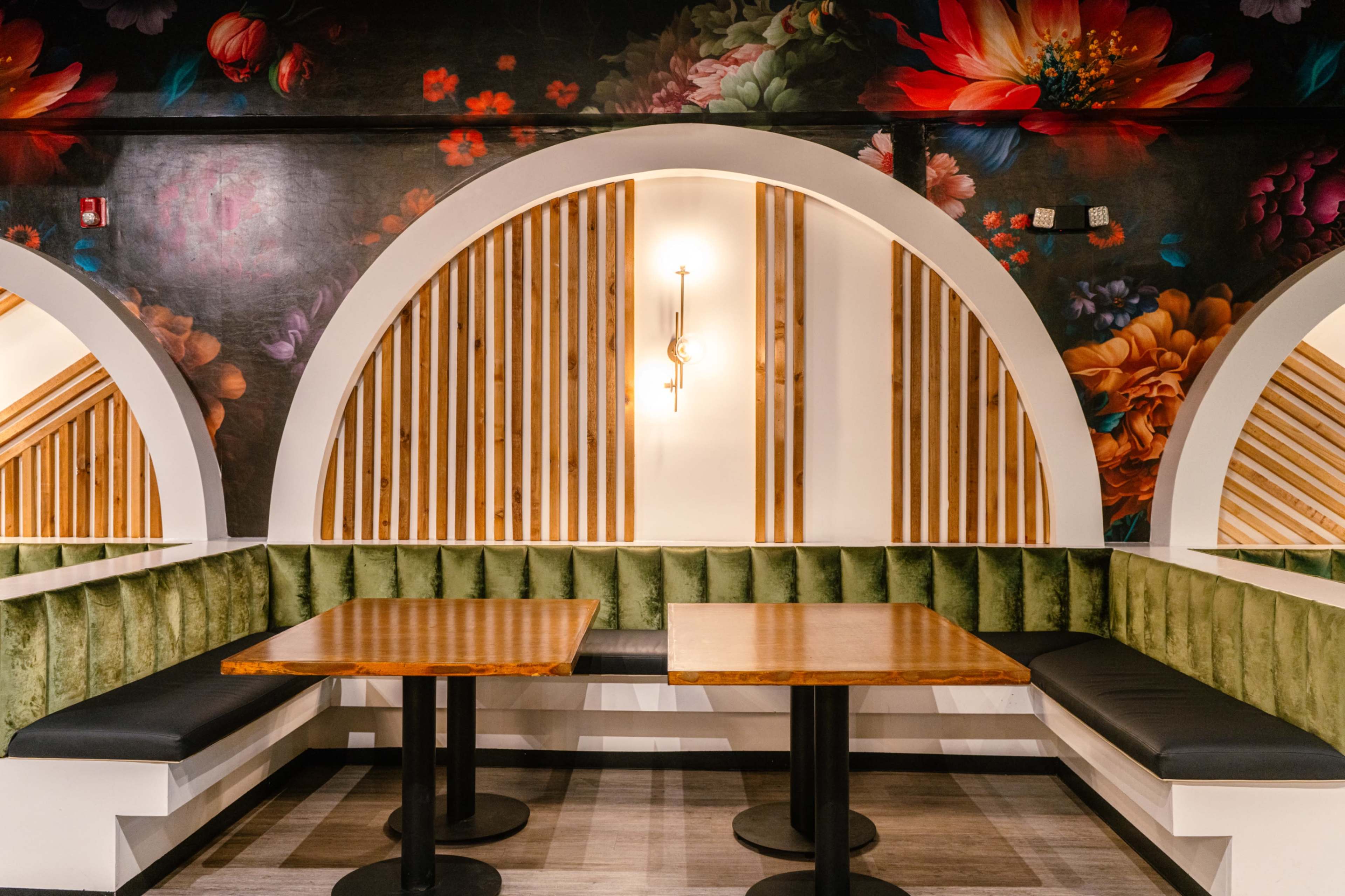 The image shows a restaurant interior featuring green velvet seating and wooden tables against a backdrop of floral wall art and curved architectural details.