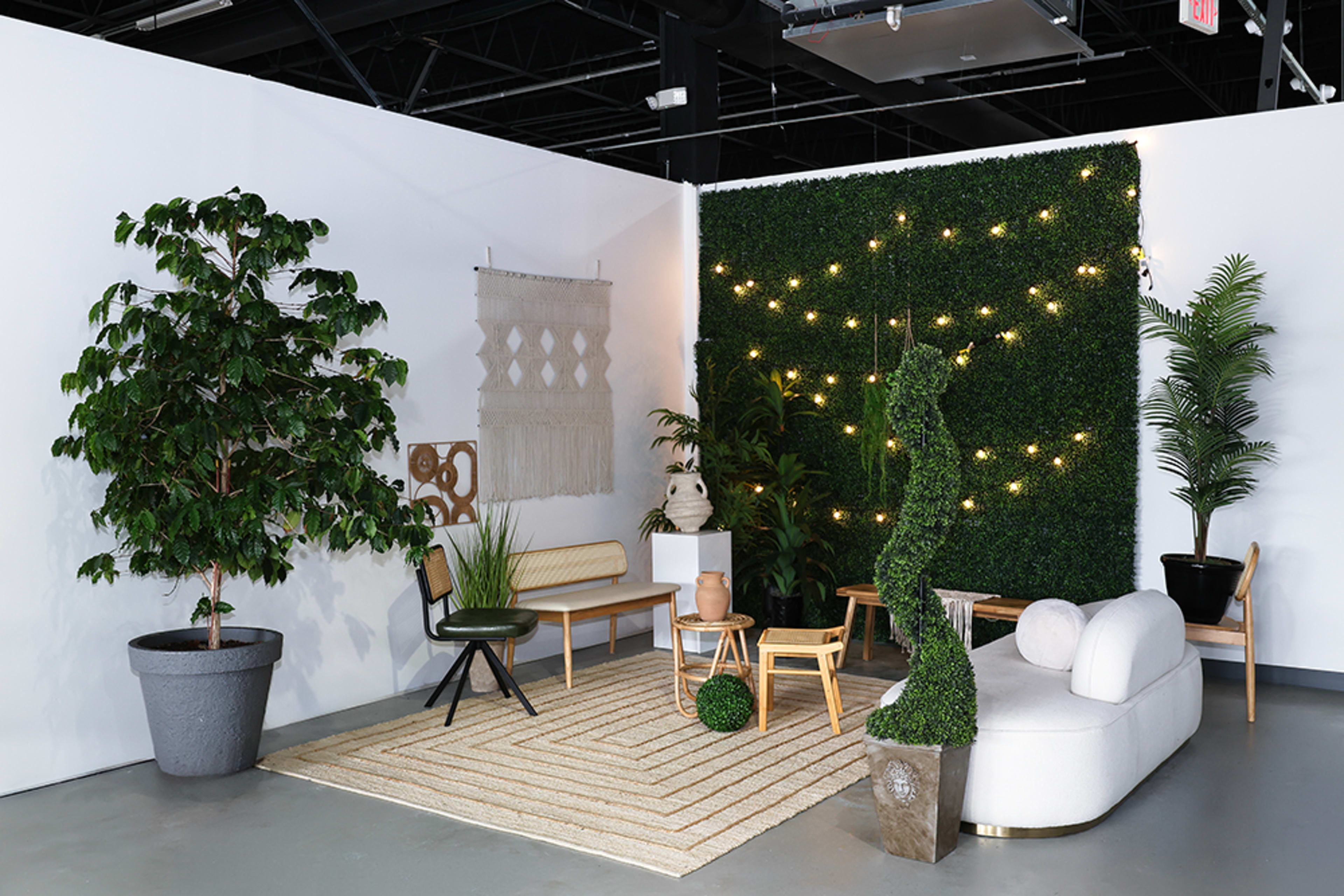 The scene features a modern interior with a seating area, including a sofa and wooden chairs, surrounded by various plants, decorative wall hangings, and string lights.