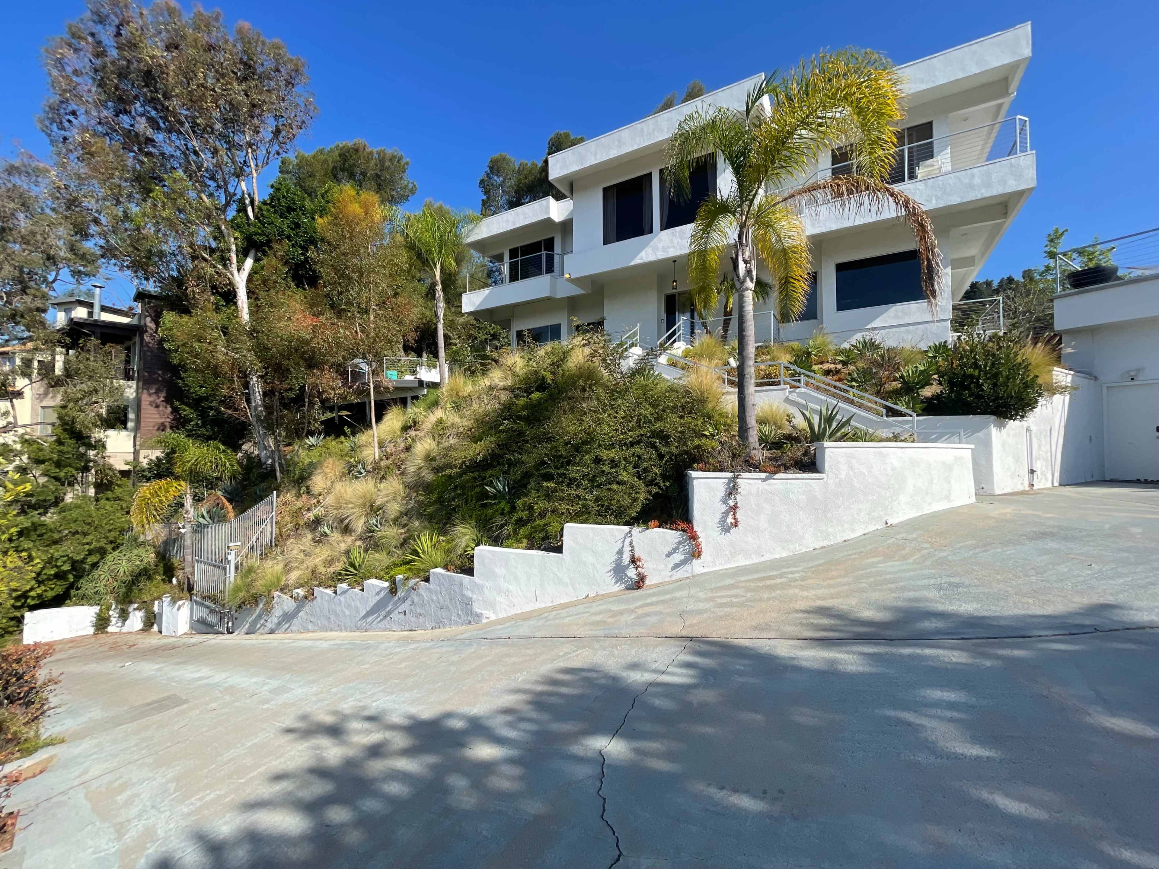 A modern white house sits on a hillside, surrounded by greenery and palm trees, with a driveway winding down to the bottom.