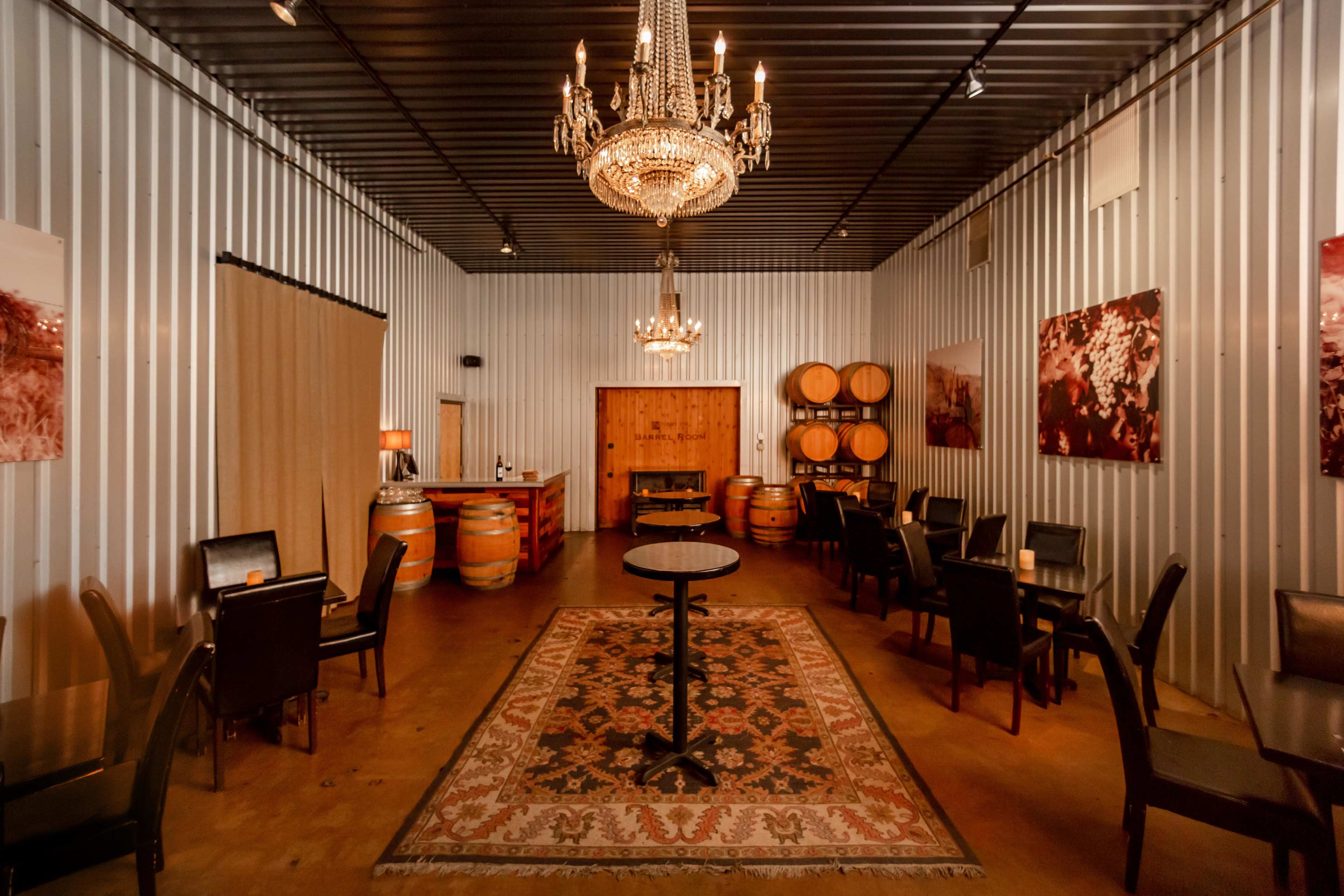 An empty room with metal walls, a chandelier, wooden barrels, a central round table, and several chairs arranged around a decorative rug.