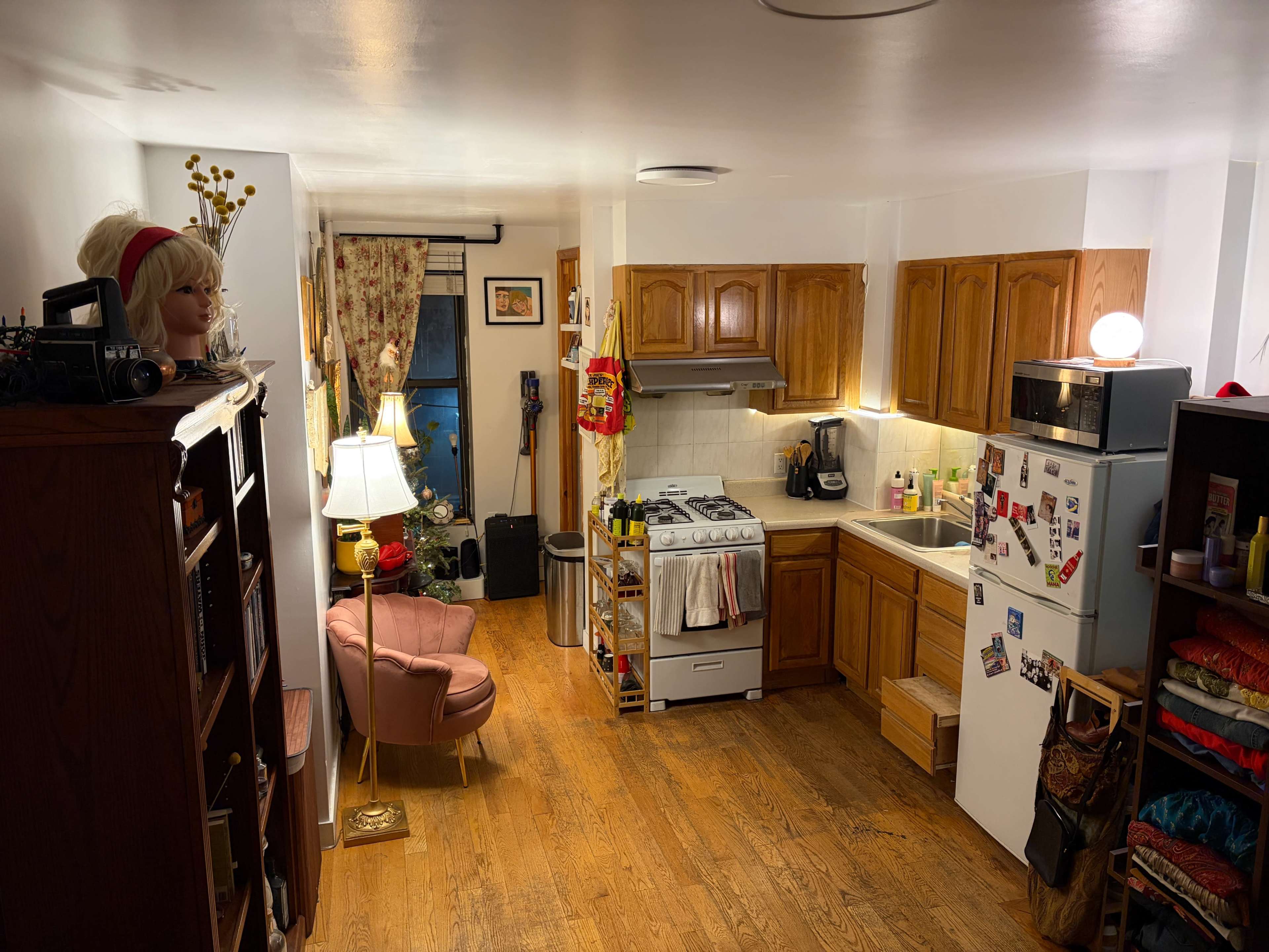 The image shows a compact kitchen and living area with wooden cabinets, a pink armchair, and various decorations, including a lamp and a faux plant.