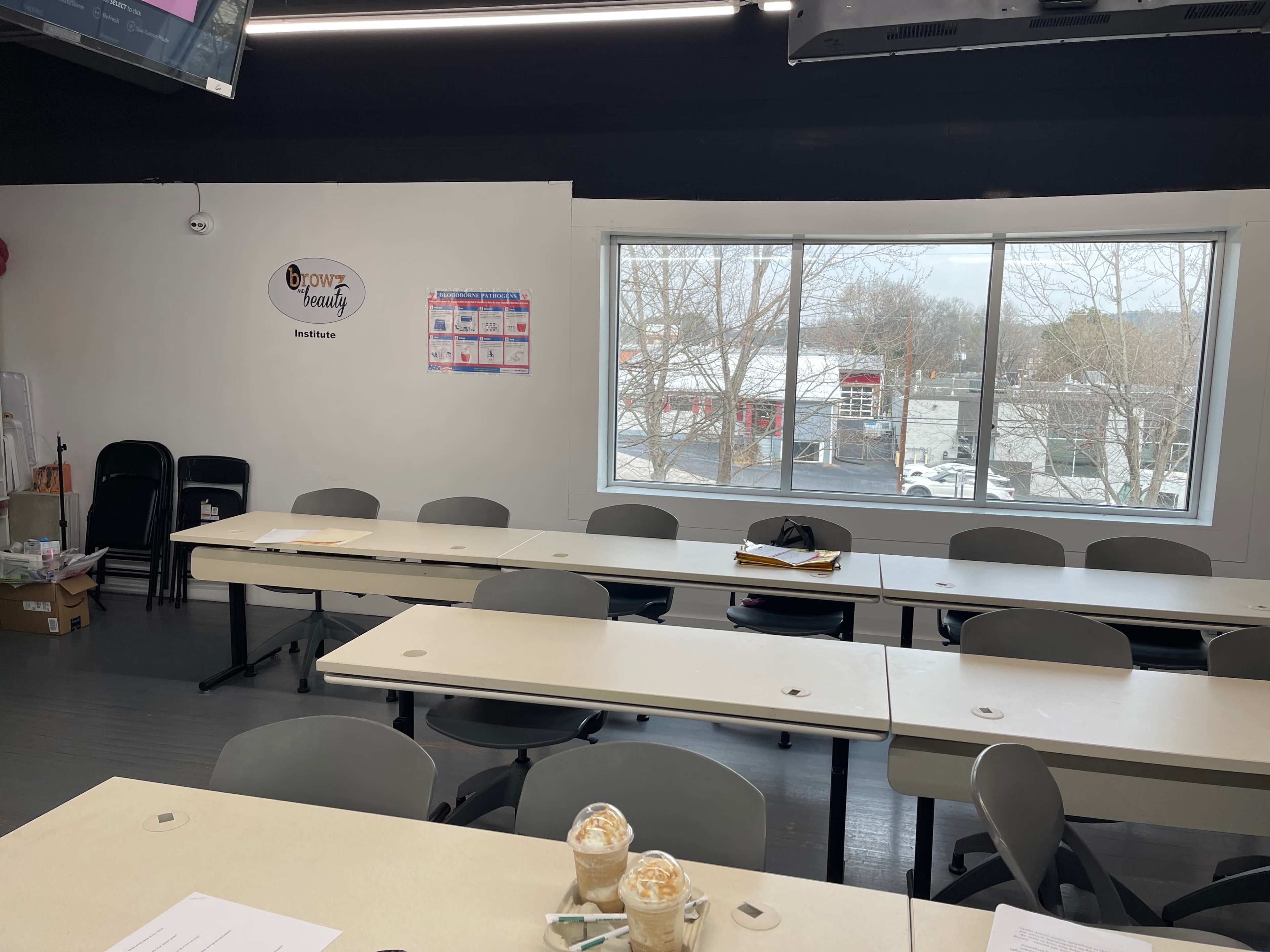 A classroom or meeting space with several tables arranged in a U-shape, facing a large window overlooking a parking lot and buildings outside.