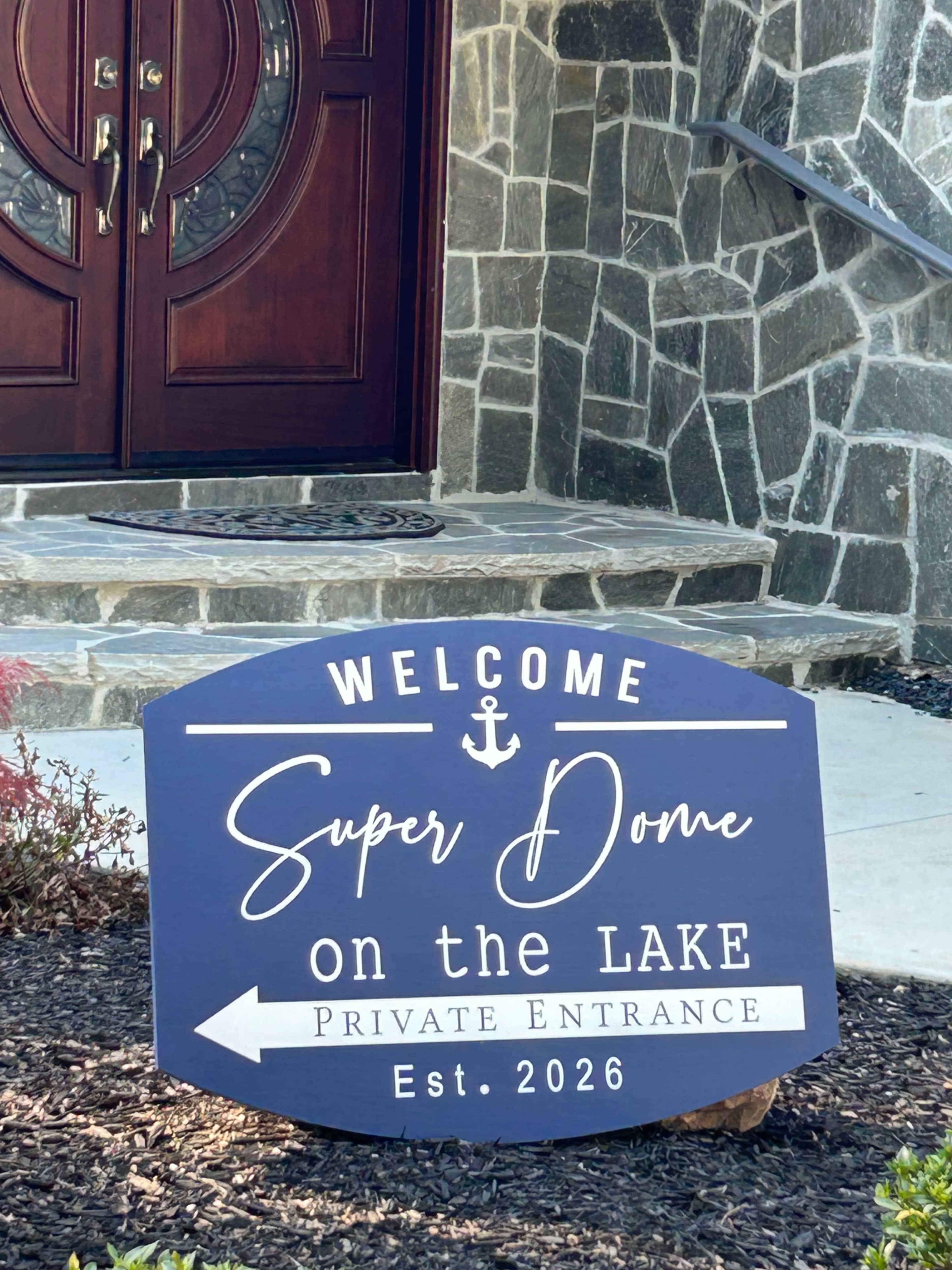 A decorative sign reading "Welcome Super Dome on the Lake Private Entrance Est. 2026" stands in front of a stone building with double doors.