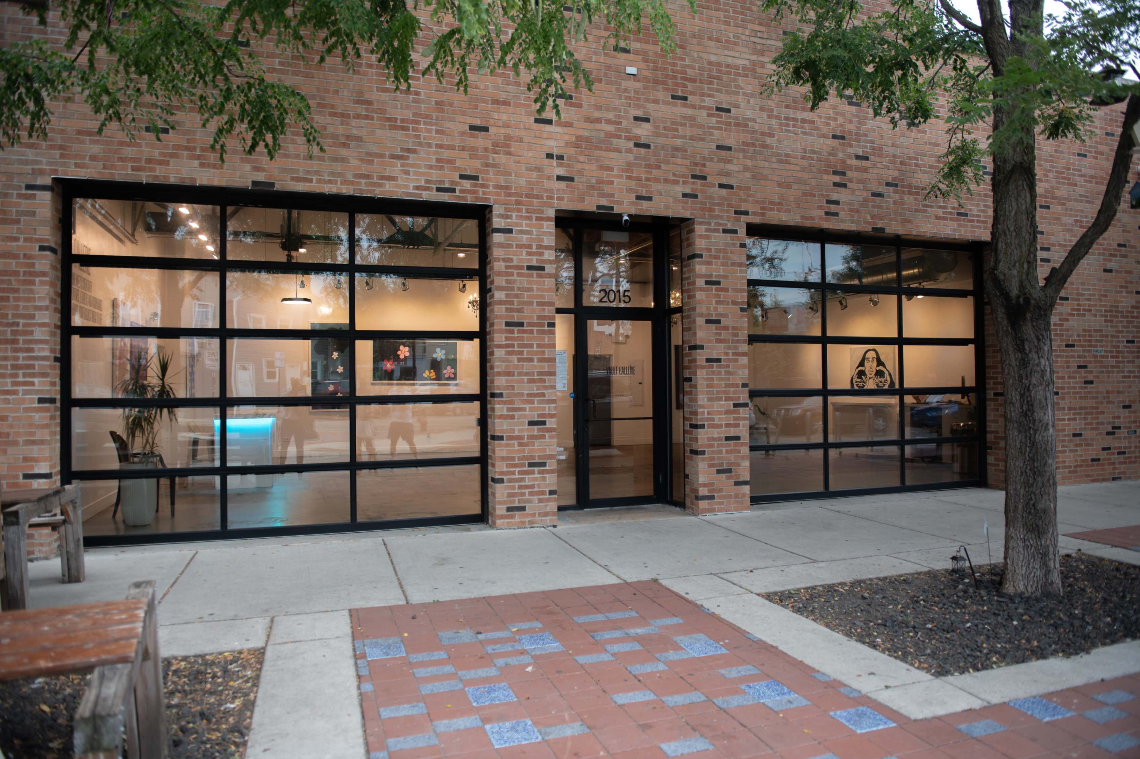 The image shows the exterior of a brick building with large glass windows and a doorway, set in a paved area with some greenery.