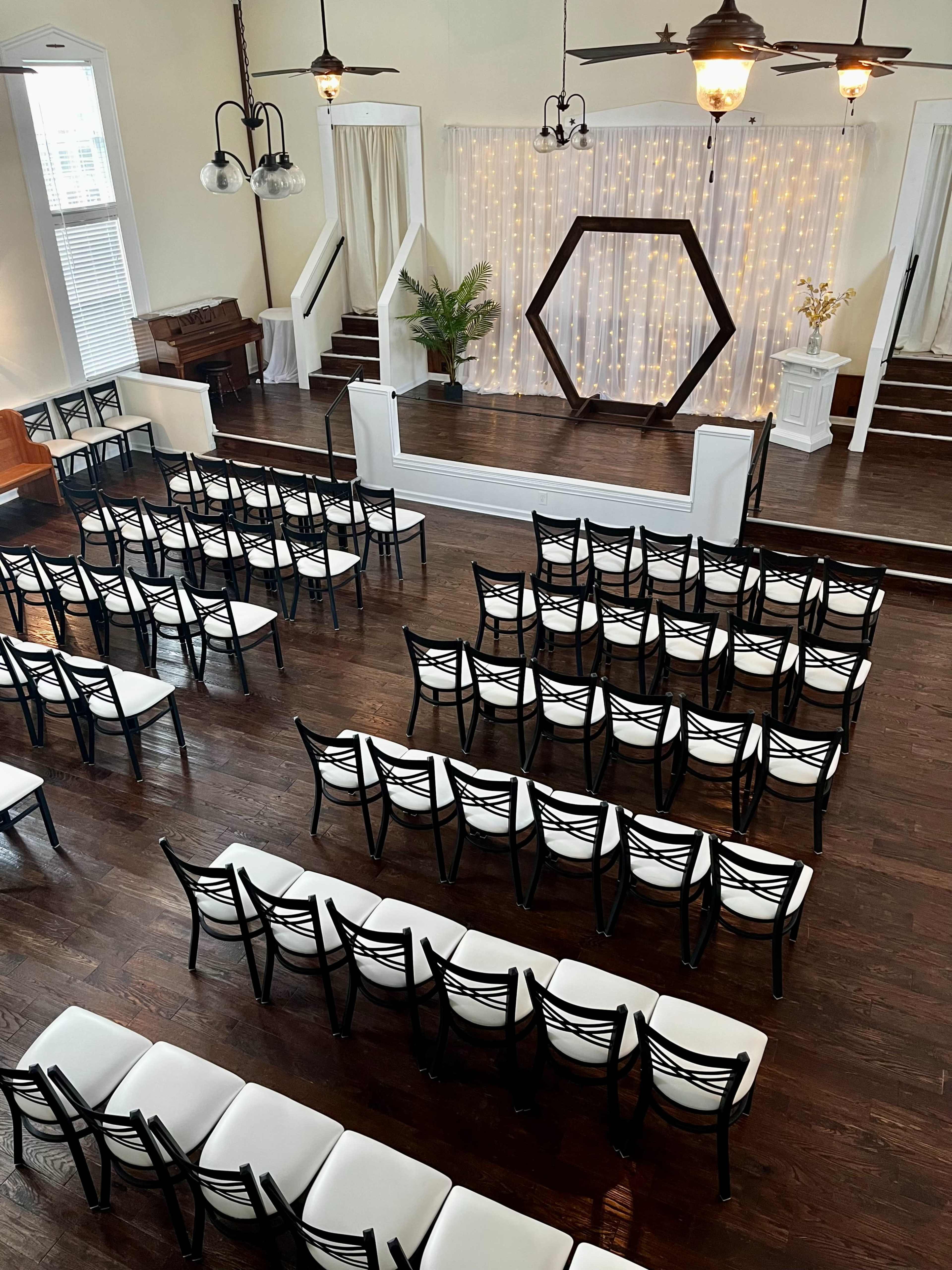The image shows an indoor venue with rows of black chairs facing a staged area that features a geometric backdrop and decorative drapes.