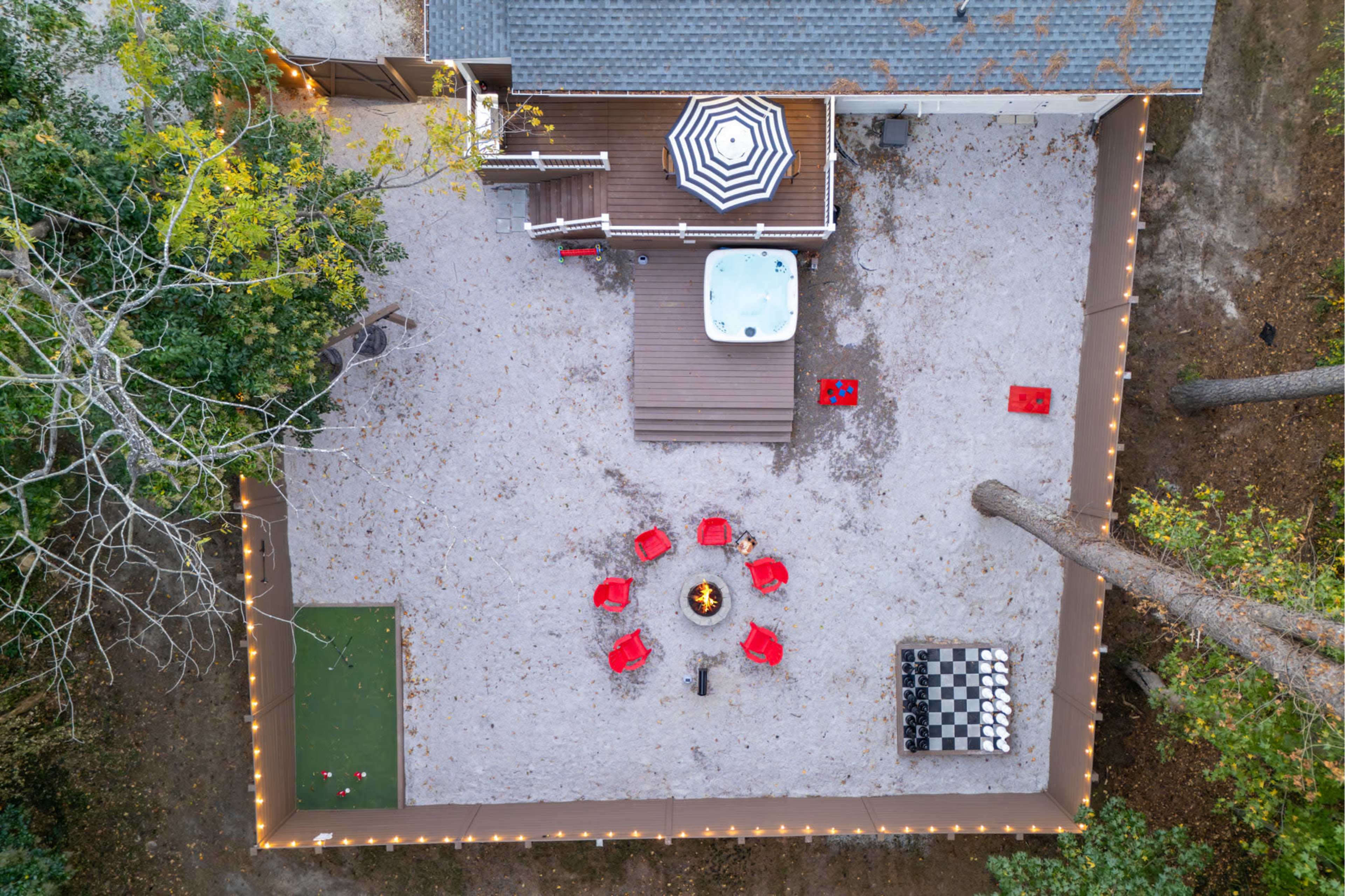 The image shows a backyard area with a hot tub, a fire pit surrounded by red chairs, a large checkers board, and a deck with an umbrella.