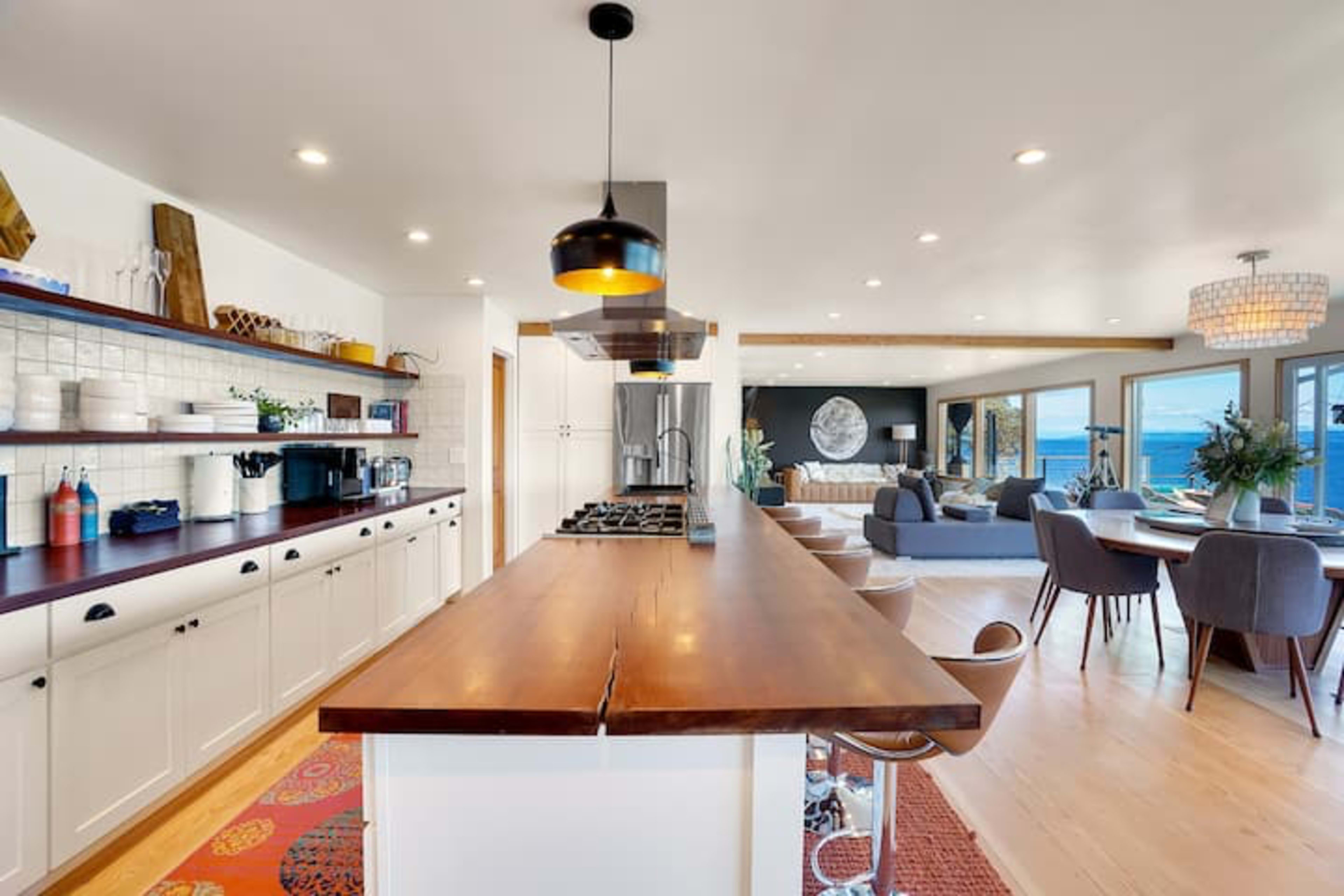 A modern kitchen with a central wooden island, featuring a stove, surrounded by a light-filled open living area that includes a dining area and seating.