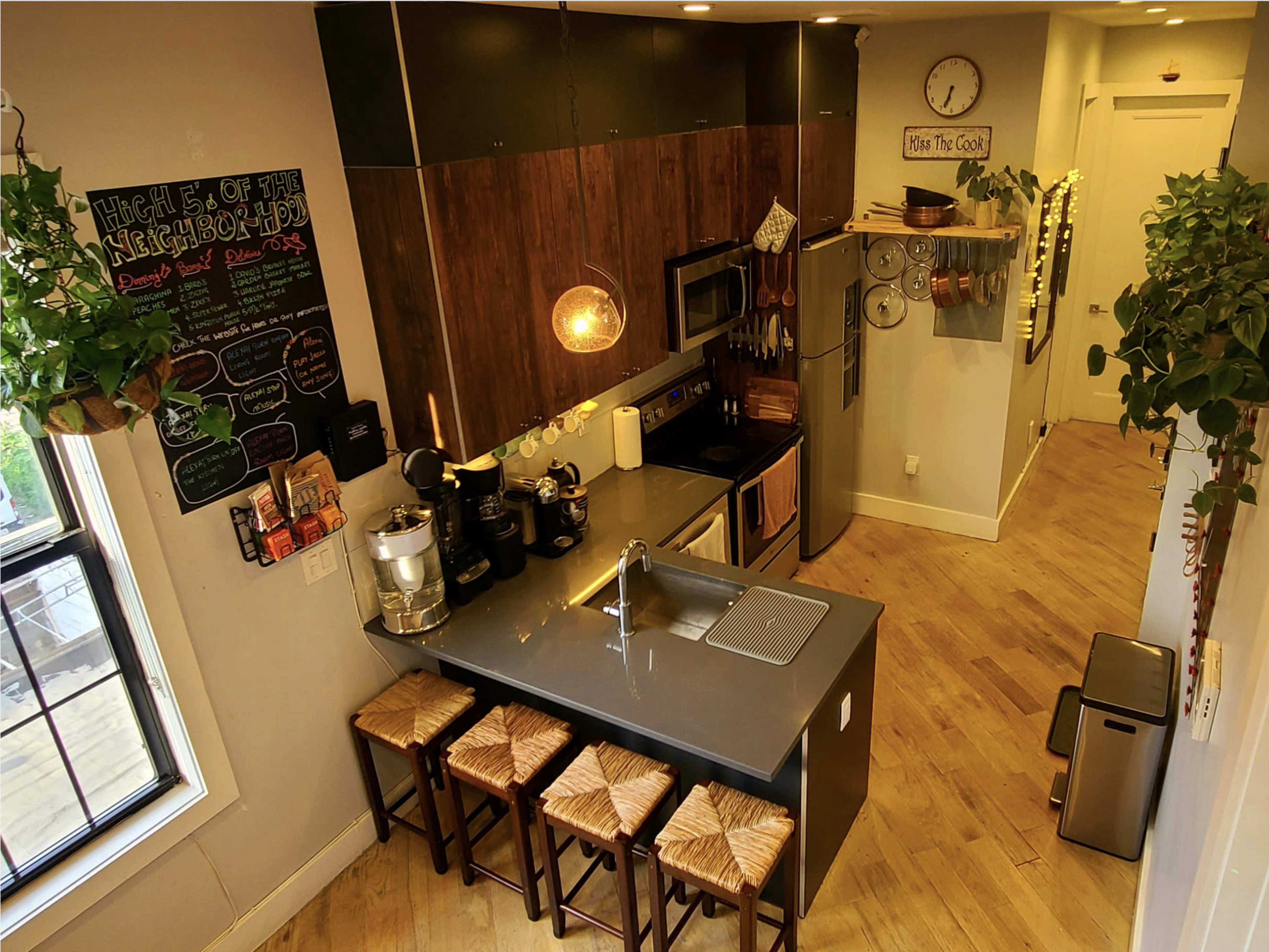 A modern kitchen with an island, equipped with stainless steel appliances, wooden cabinetry, and three stools, surrounded by plants.