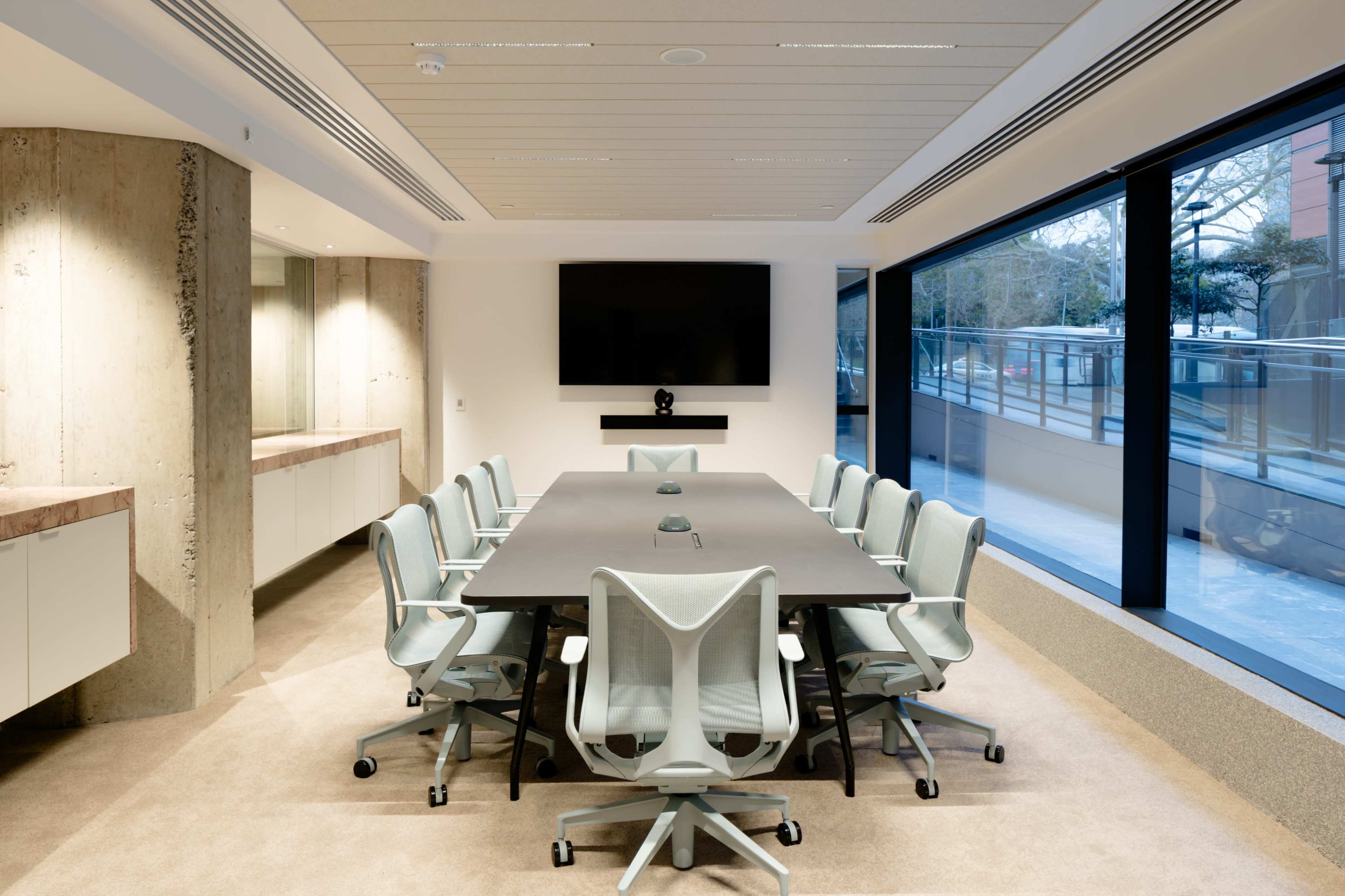 A modern conference room features a large rectangular table surrounded by ergonomic chairs, with a large screen mounted on the wall.