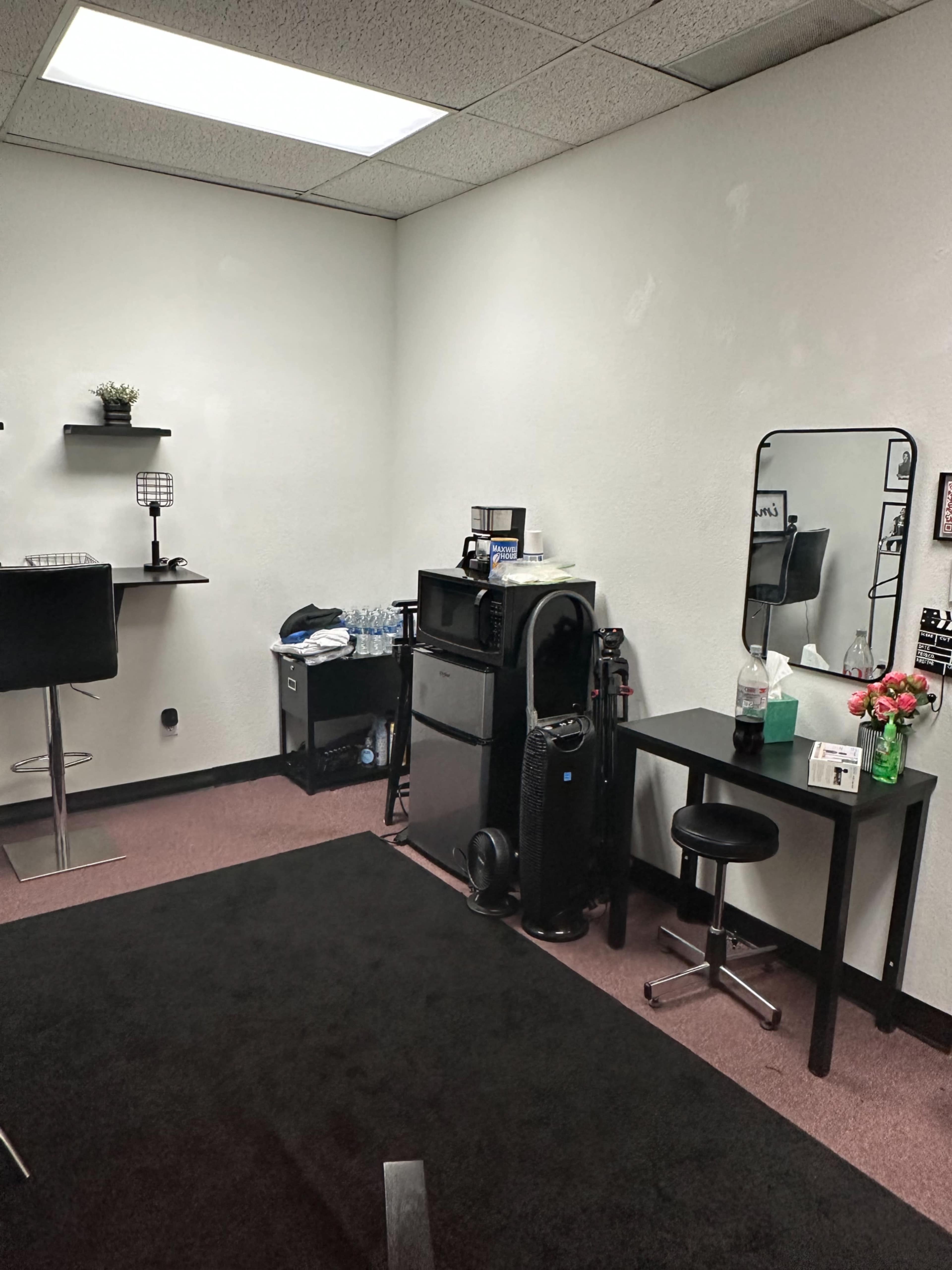 A small, sparsely furnished office space featuring a black carpet, a table with a mirror, a mini fridge, and a bar stool.