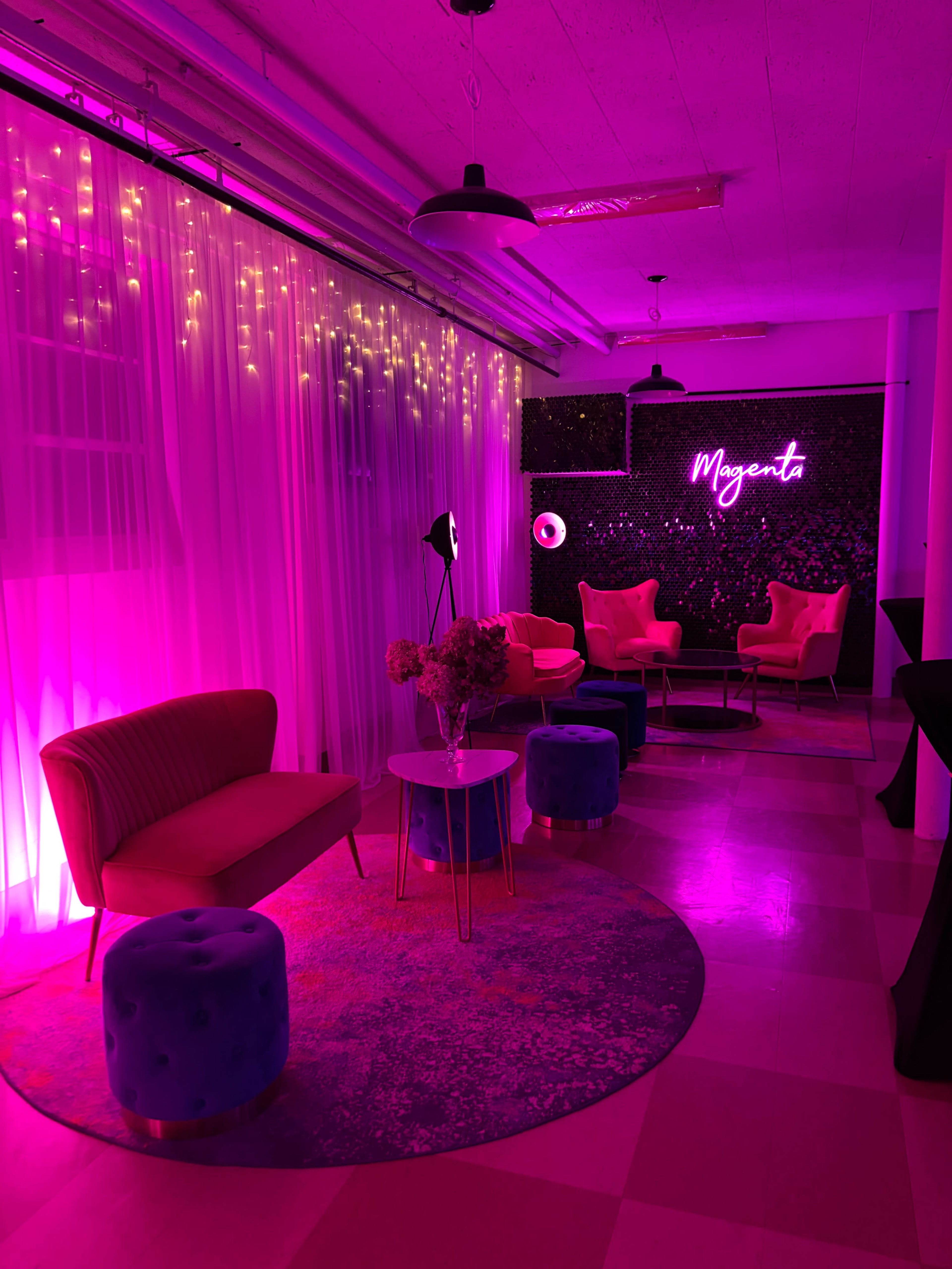 The image shows a brightly lit lounge area with pink lighting, featuring colorful seating, a decorative rug, and a neon sign that reads "Magent."