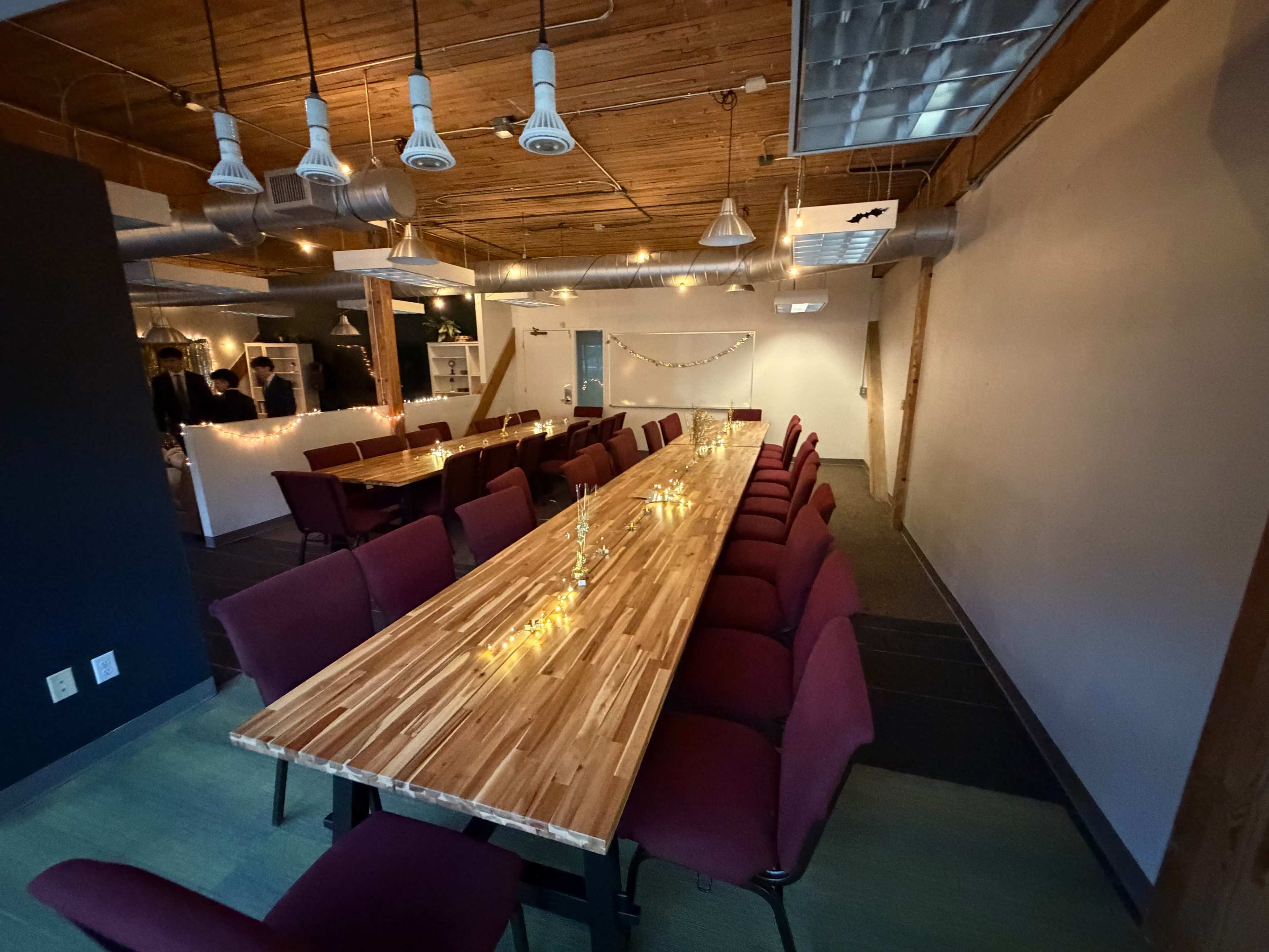 The image shows a long wooden conference table surrounded by burgundy chairs in a well-lit room with wooden ceilings and decorative string lights.