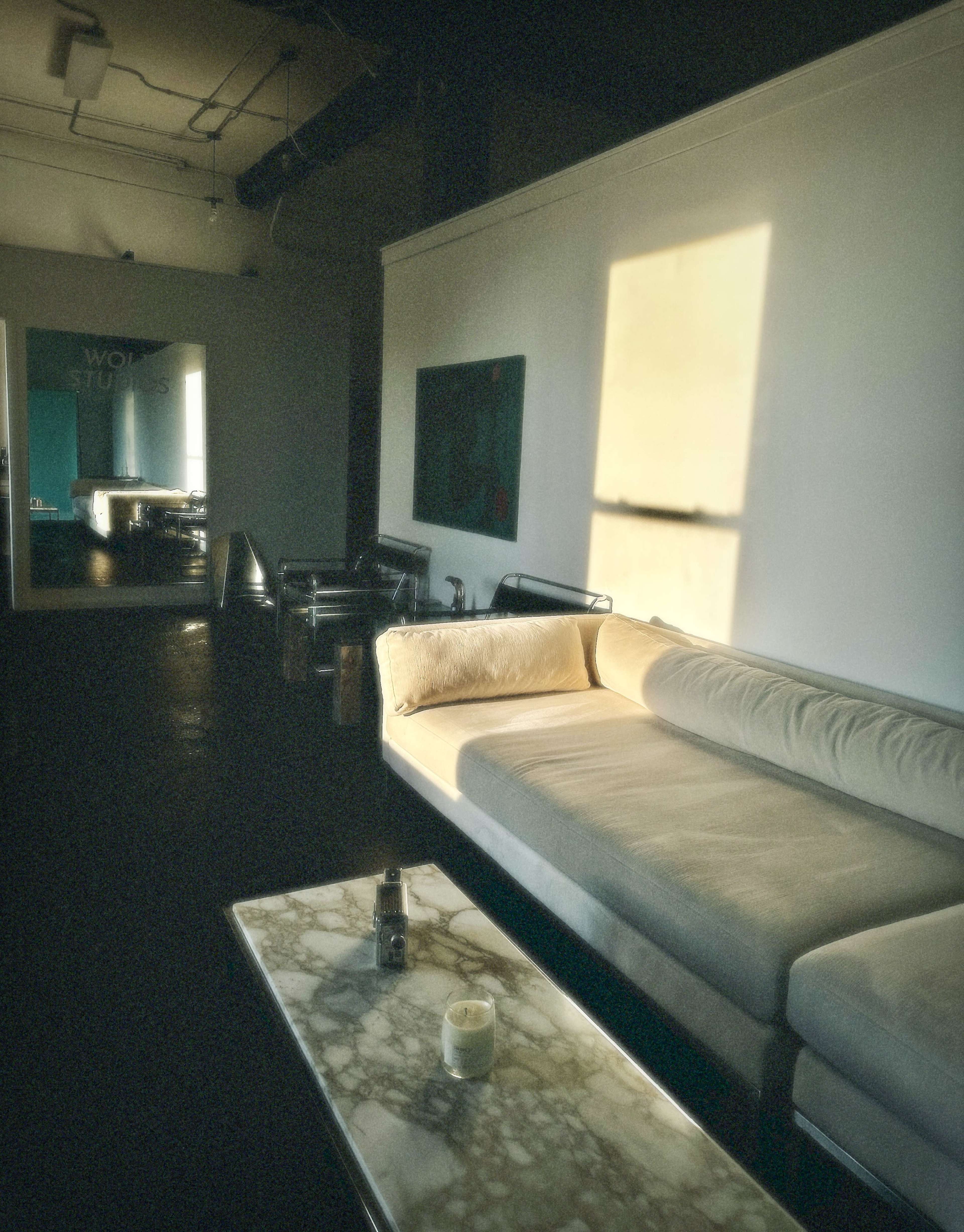 A sunlit living space features a light-colored sofa and a marble coffee table, with reflection visible in a mirror on the wall.