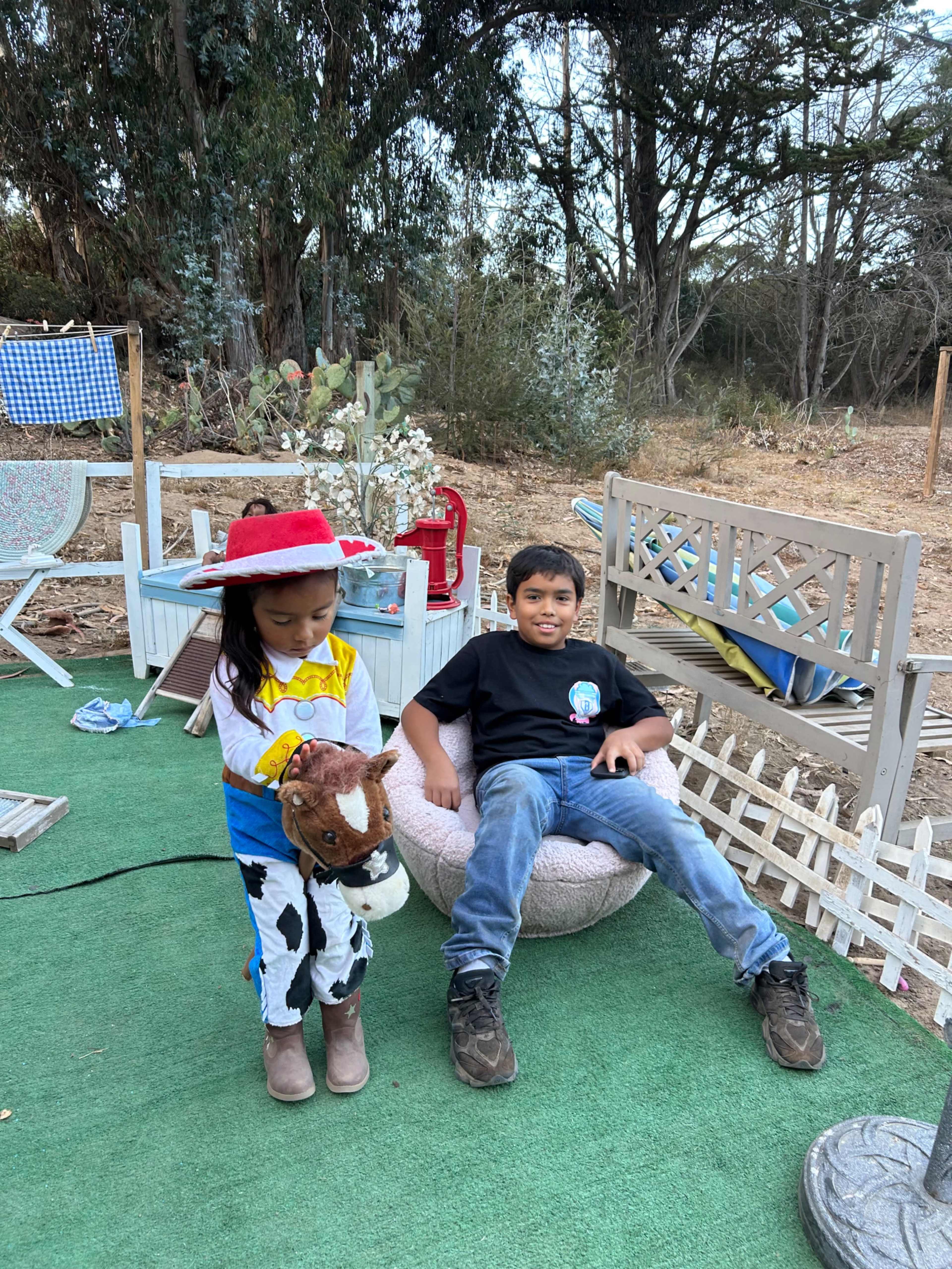 A young girl dressed as a cowboy holds a toy horse while a boy sits on a bean bag chair in a rustic outdoor setting.