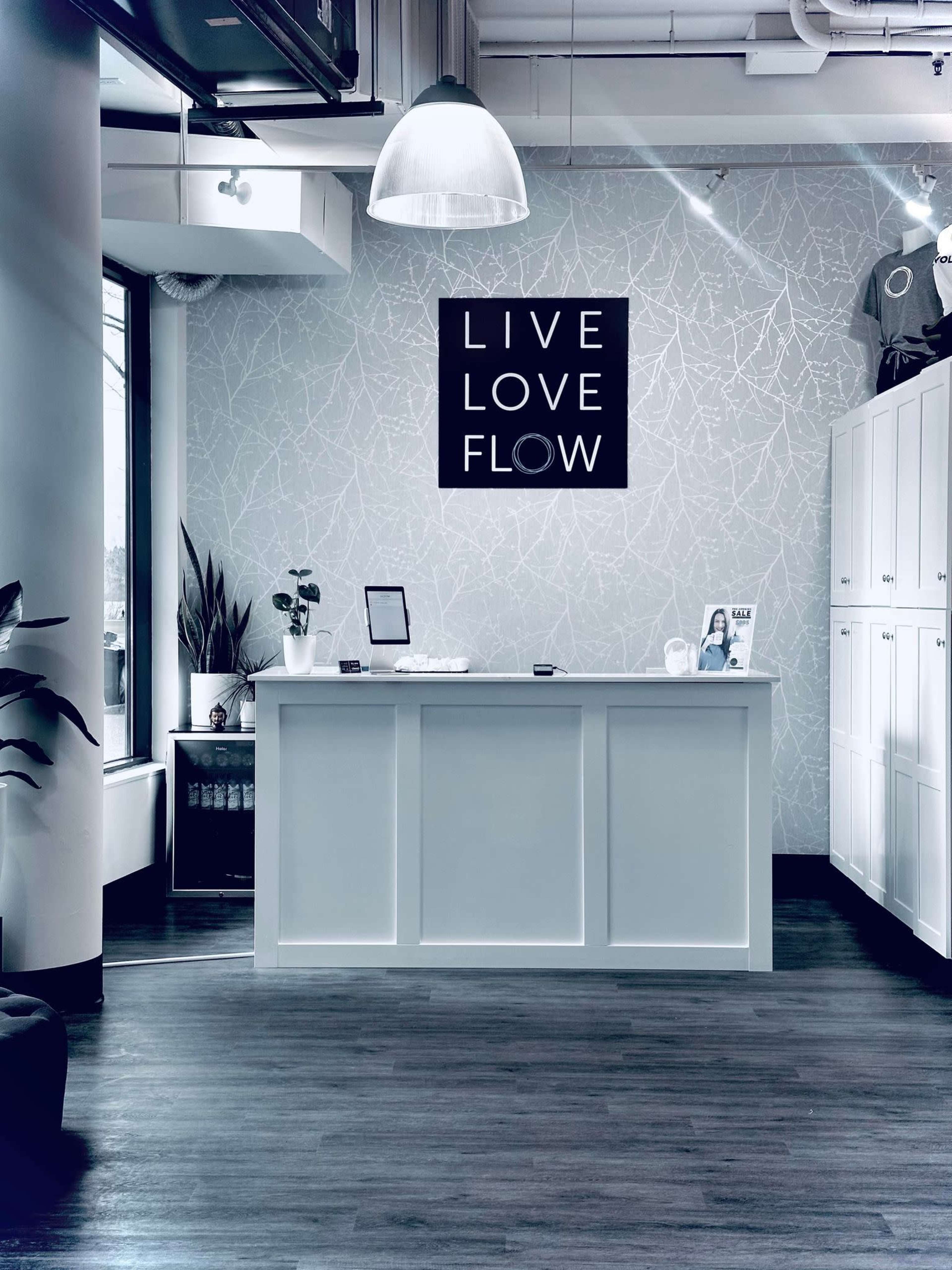 The image shows a reception area with a white desk, minimalist decor, and a framed sign that reads "LIVE LOVE FLOW" on the wall.