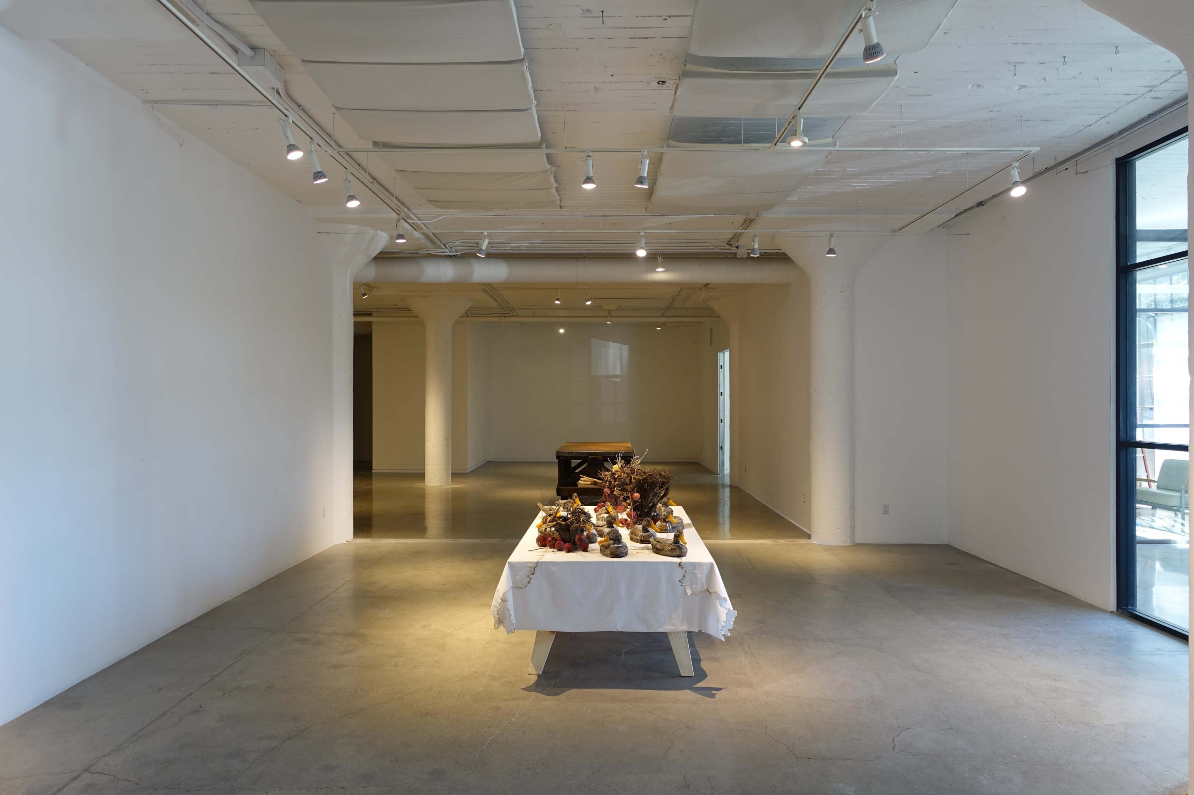The image shows a spacious, minimalist interior with a table covered in a white cloth displaying decorative items, surrounded by open spaces and soft lighting.