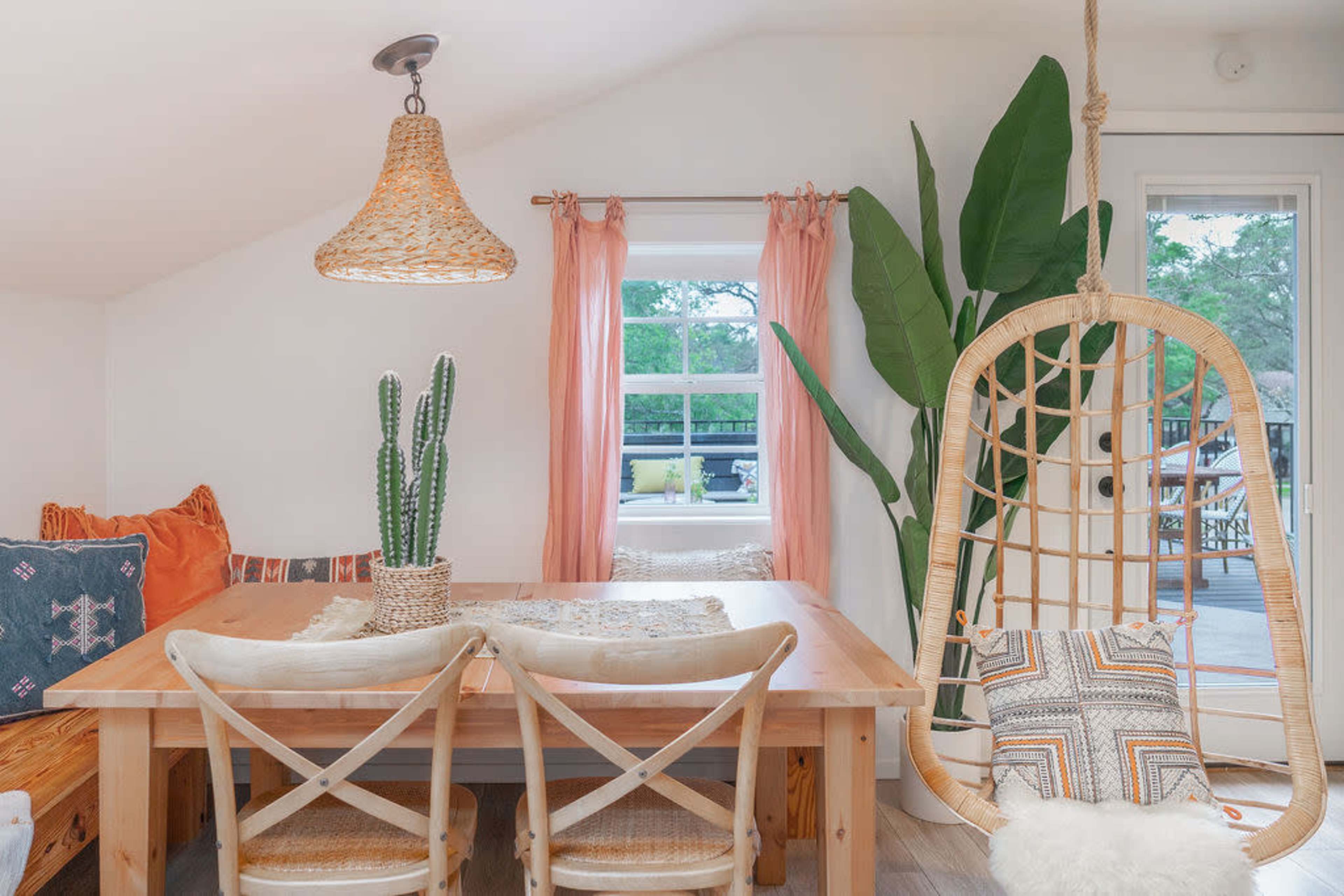 A light and airy dining area features a wooden table surrounded by chairs, a hanging rattan light fixture, a swing chair, and decorative plants.