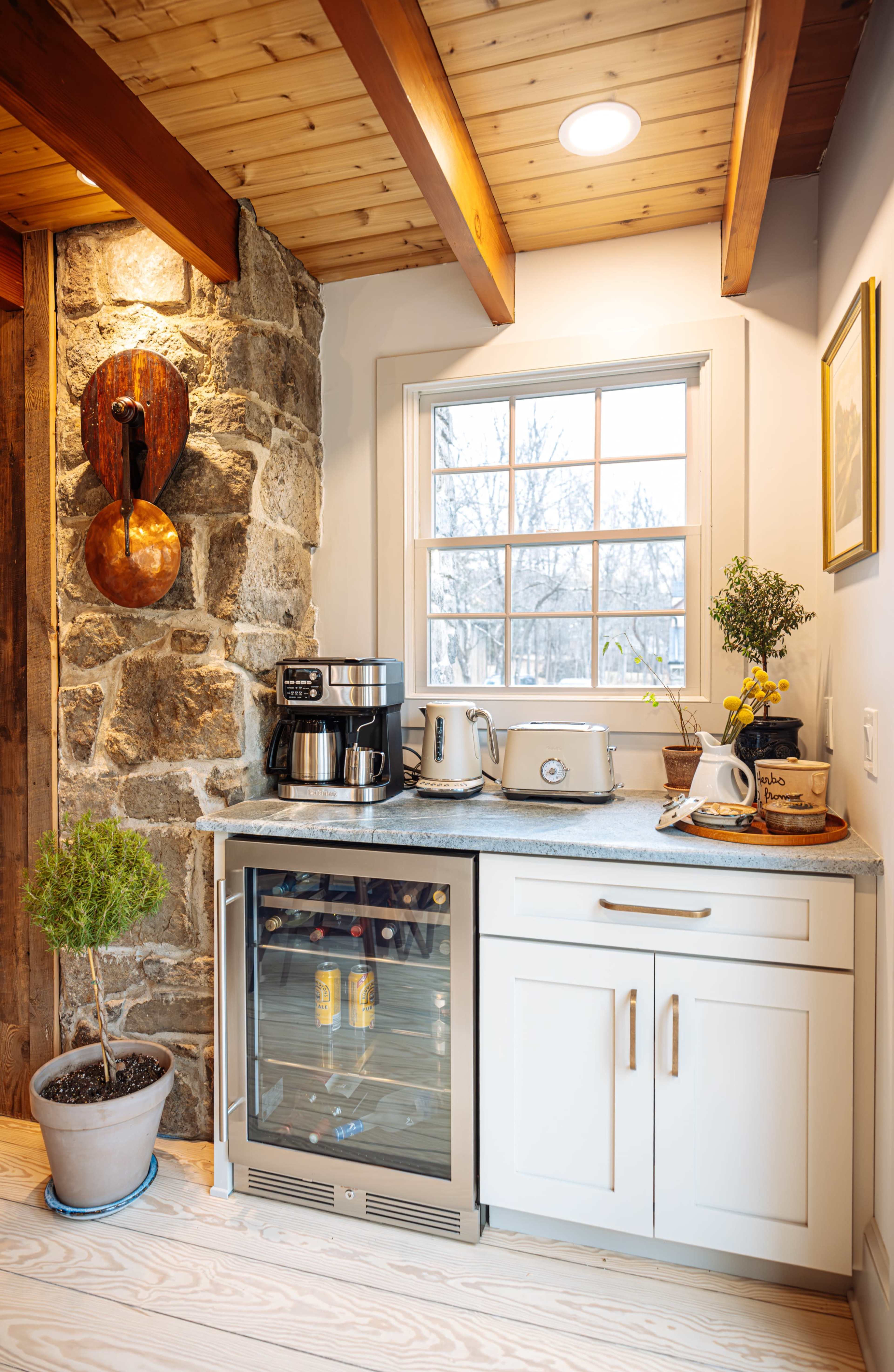 The image features a corner kitchen with a stone wall, a small fridge, coffee maker, toaster, and a window with a potted plant on the countertop.