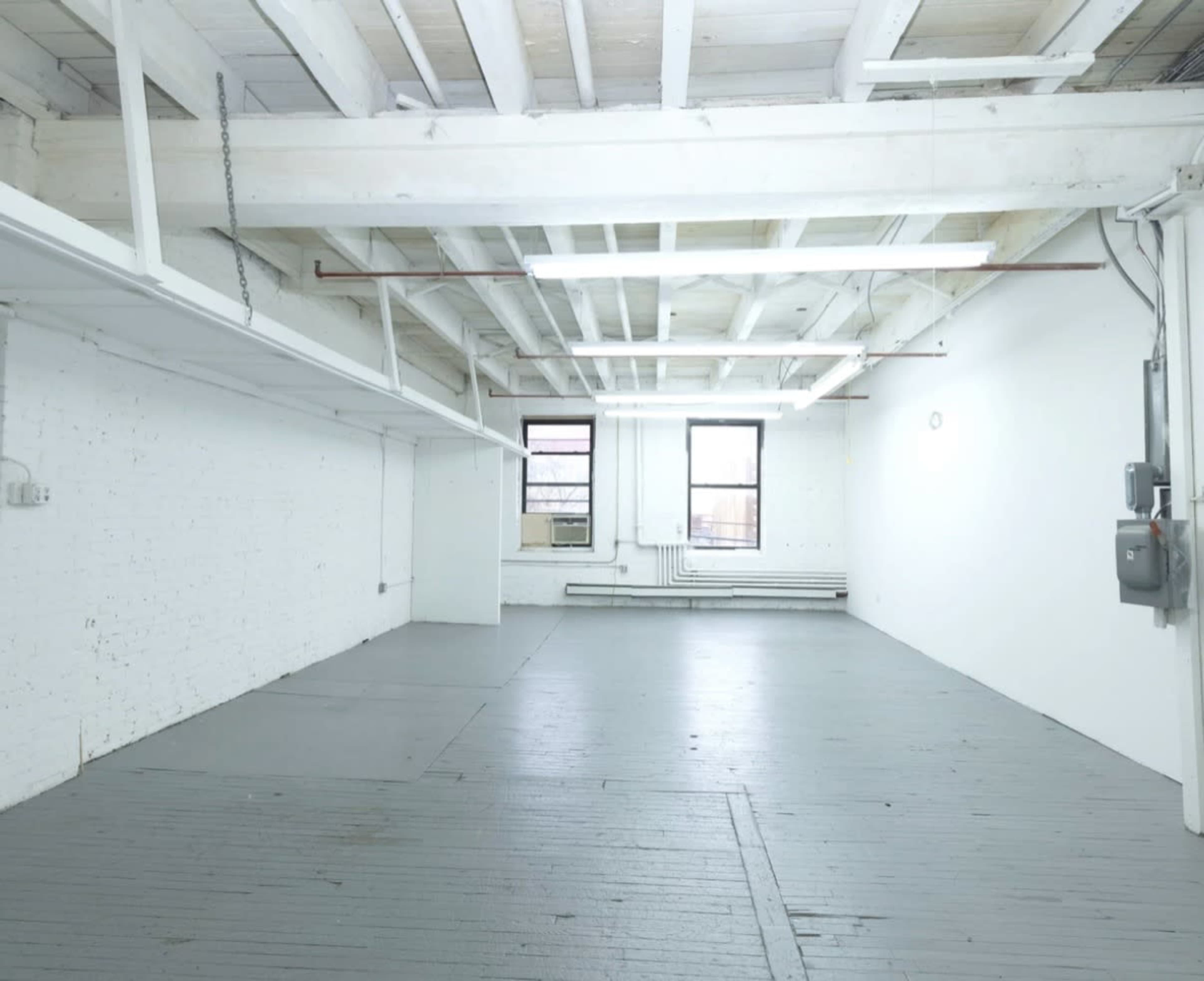 The image shows an empty, well-lit room with white walls, exposed beams, and a gray wooden floor.