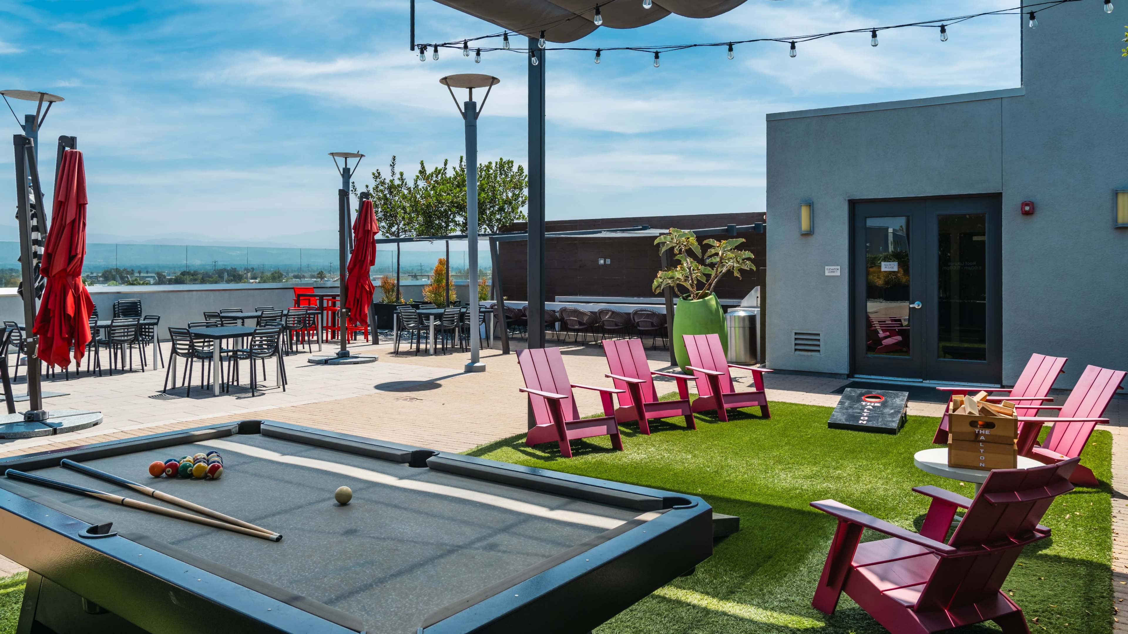 The image shows a rooftop terrace with a billiard table, several pink lounge chairs, outdoor tables, and red umbrellas overlooking a scenic view.