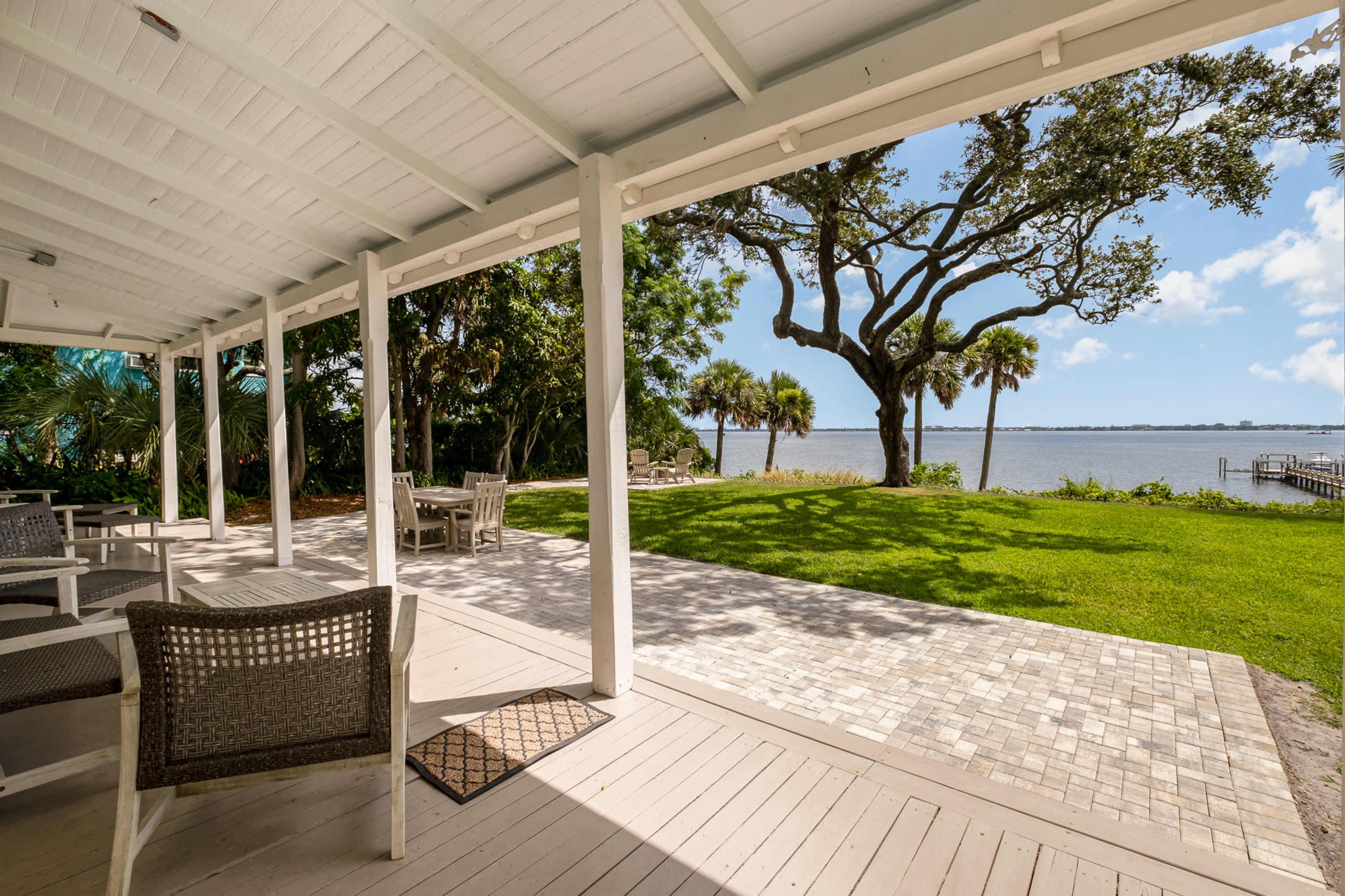 A covered patio area with seating, overlooking a lawn and a body of water with trees in the background.