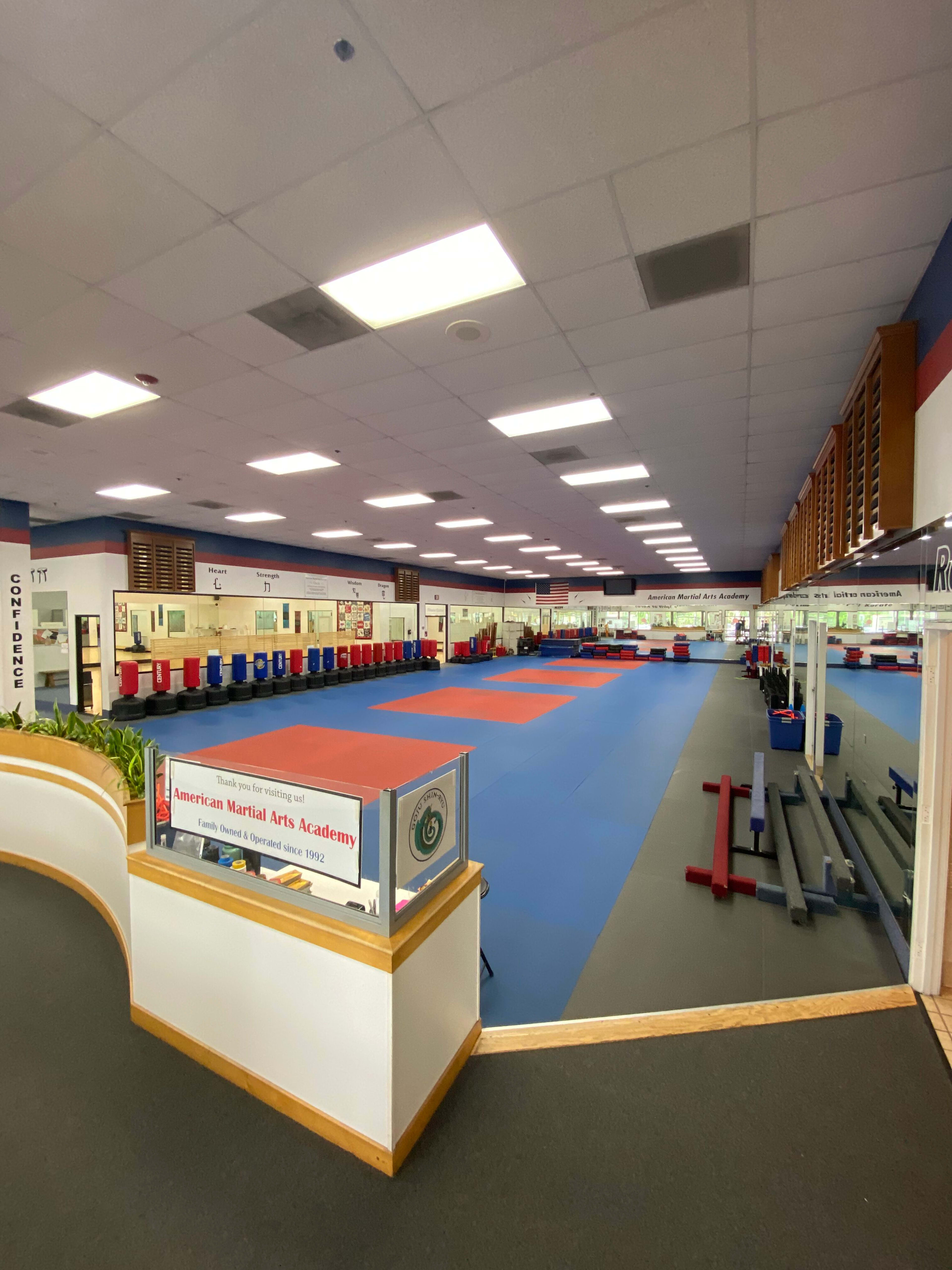 The image shows a spacious martial arts training facility with colorful mats, lined with benches and training equipment along the walls.