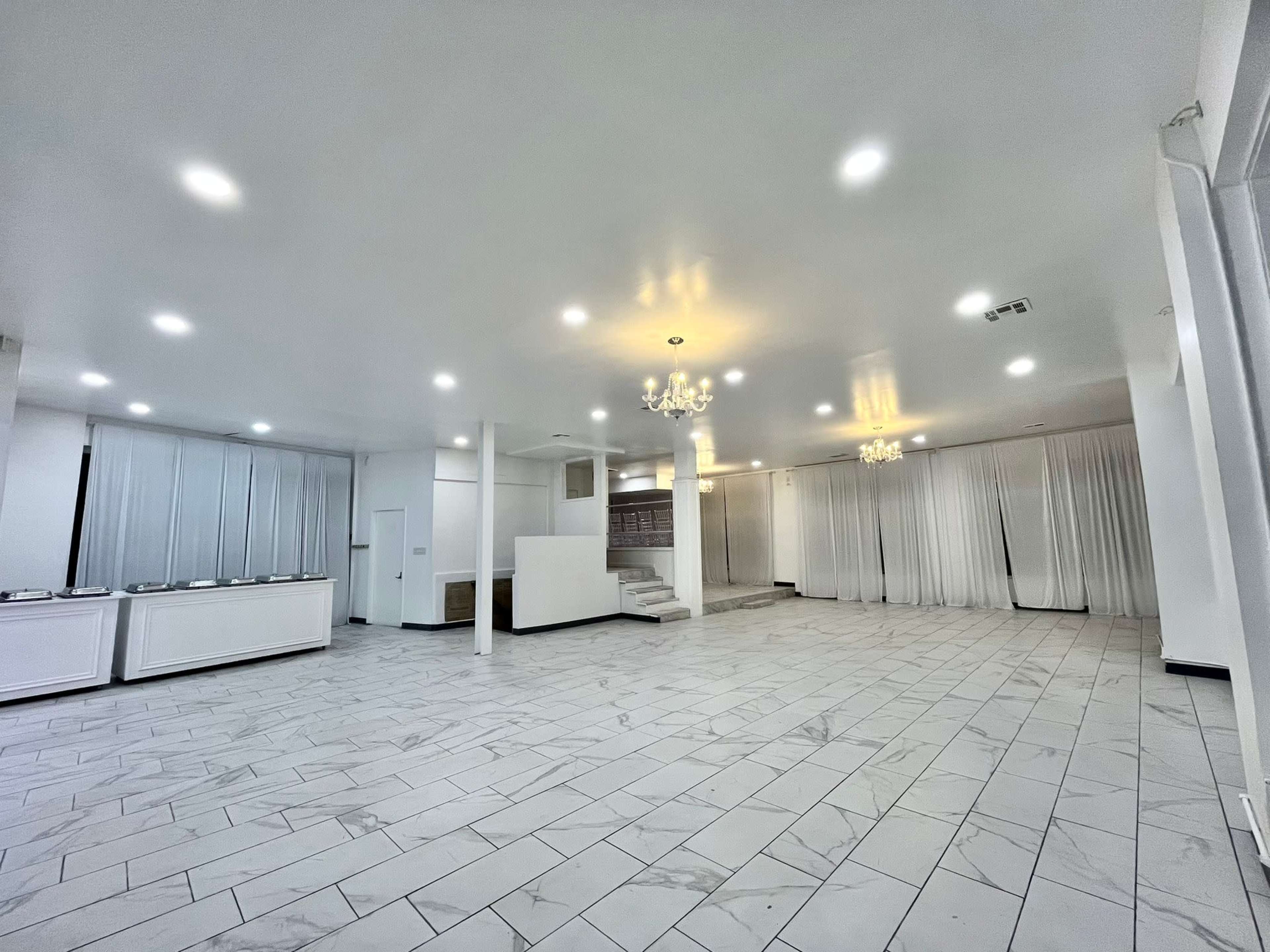 A spacious, empty interior room with white walls, tiled flooring, and multiple chandeliers hanging from the ceiling.