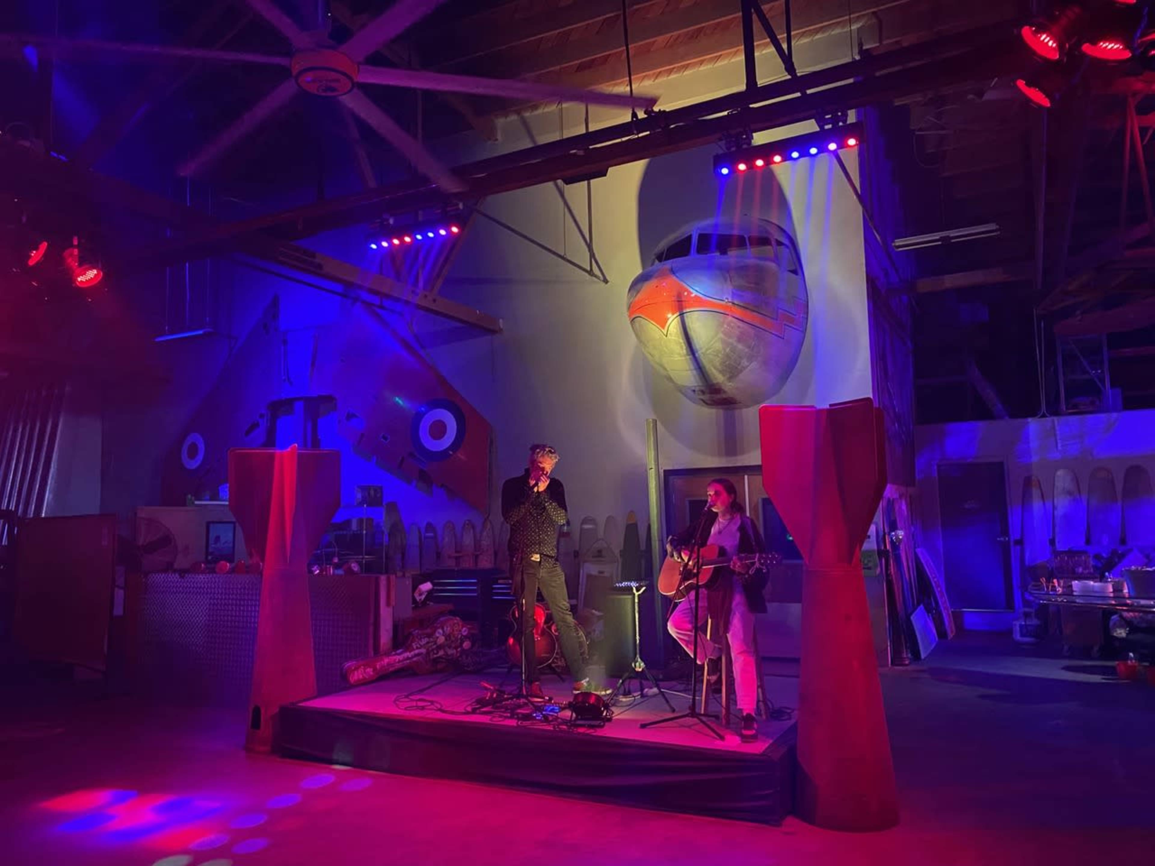 A singer and an acoustic guitarist perform on a small stage in a colorful, industrial space decorated with airplane-themed artwork.