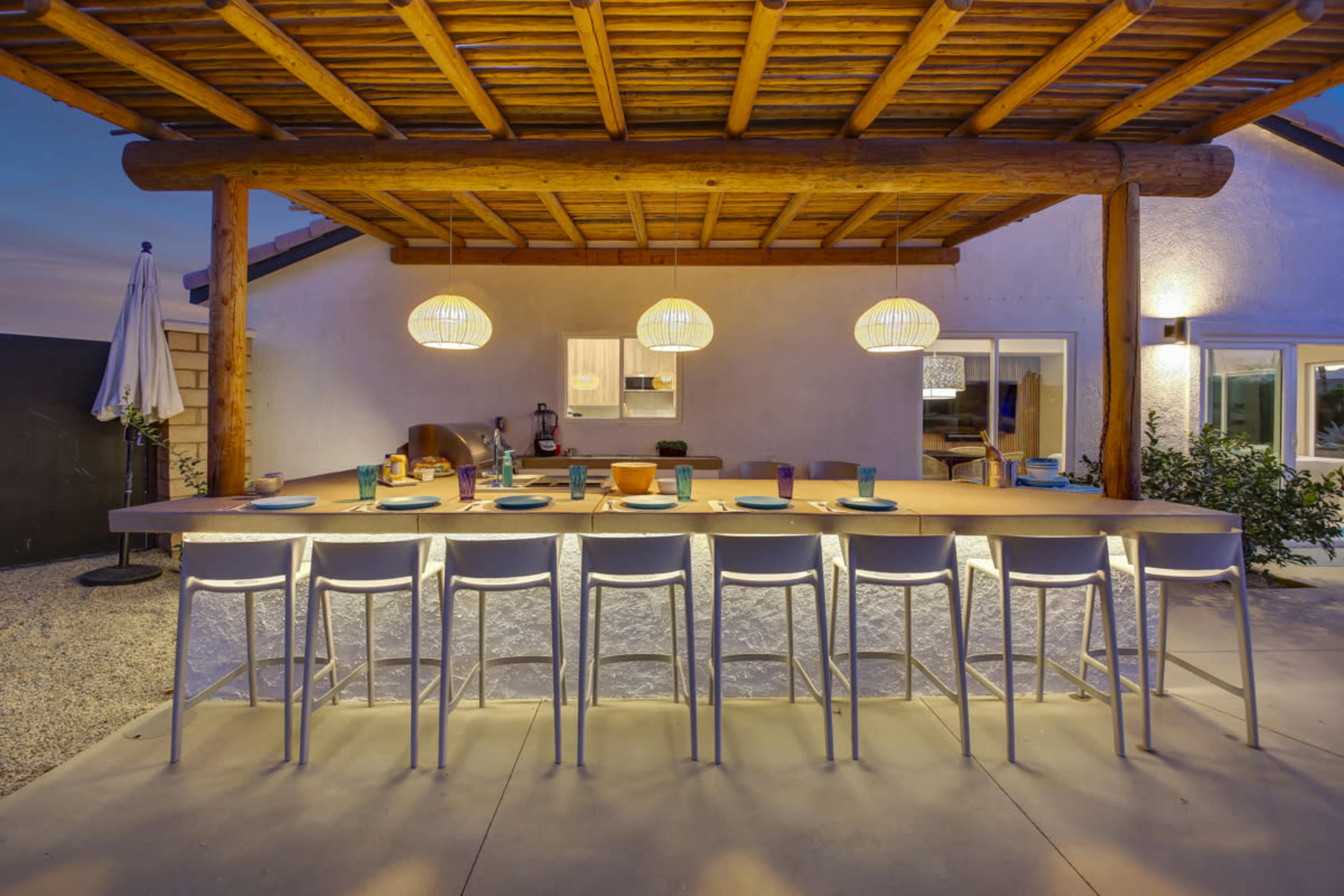 A patio area with a long illuminated bar counter and three pendant lights hanging above it, surrounded by chairs and featuring a backdrop of a home and garden.