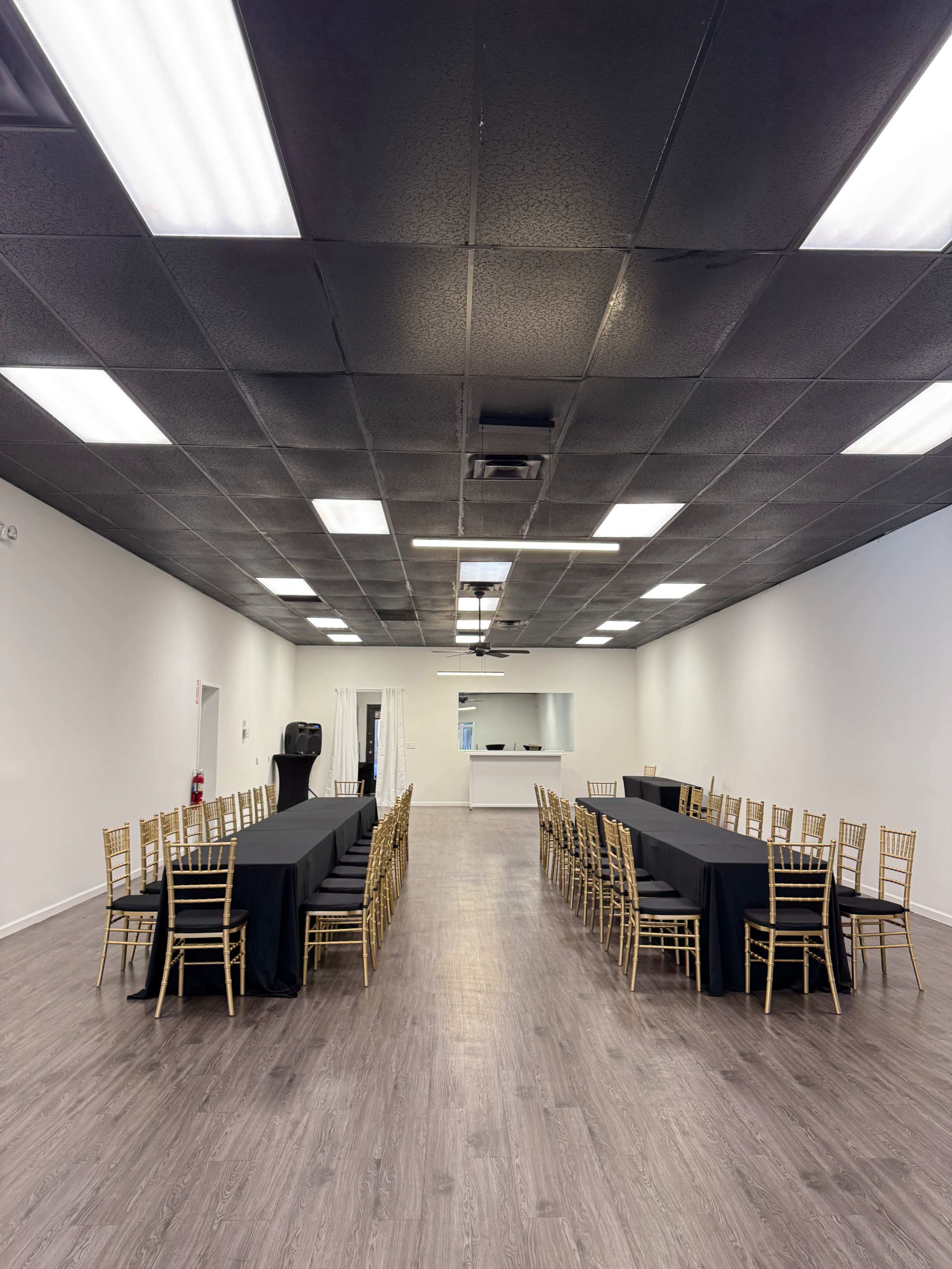 The image shows a spacious event room with two long tables covered in black linens, surrounded by gold chairs, and a simple service area at the far end.