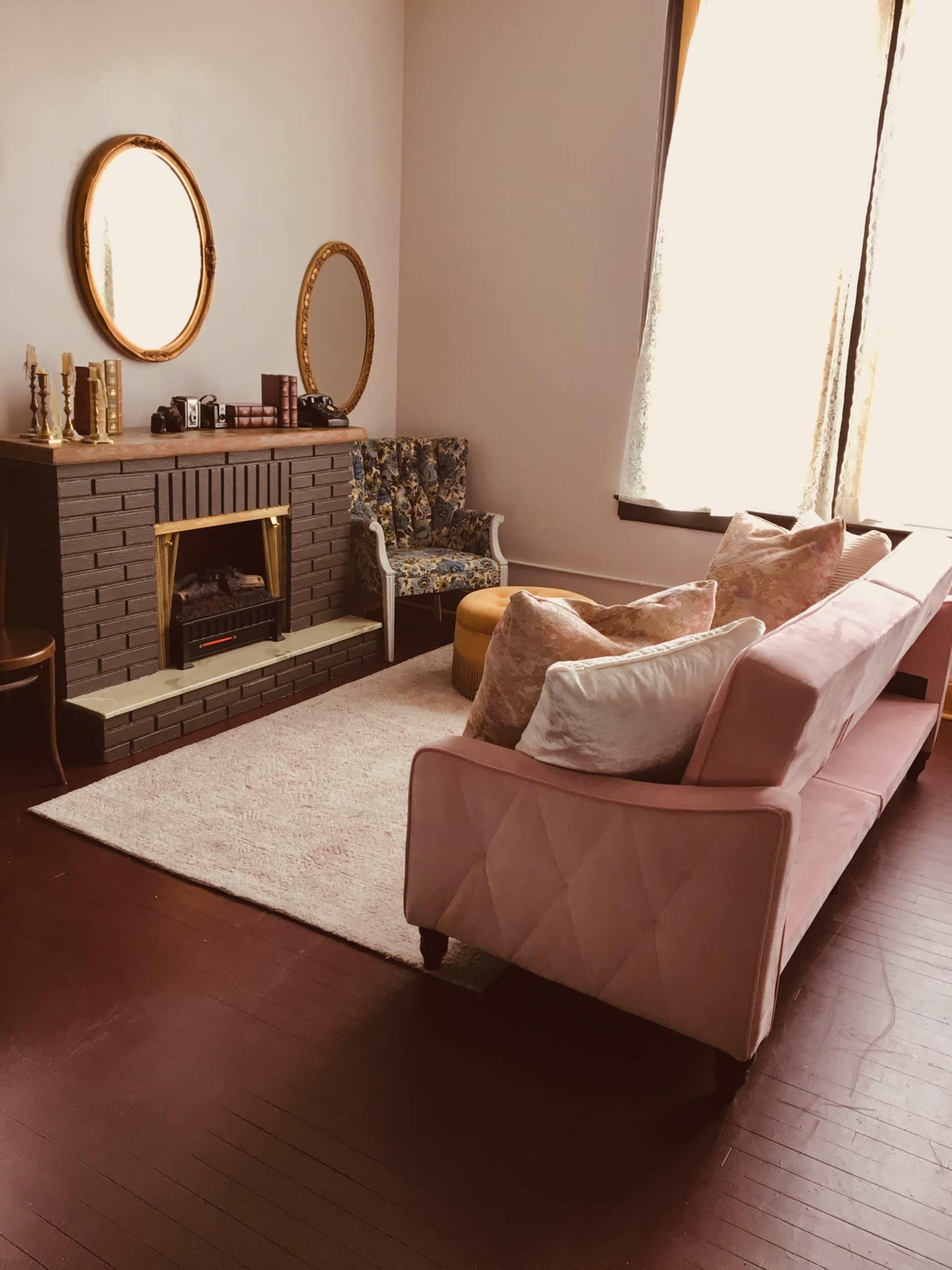 A cozy living room with a pink couch, a patterned armchair, a gold-framed mirror, and a fireplace surrounded by decorative items.