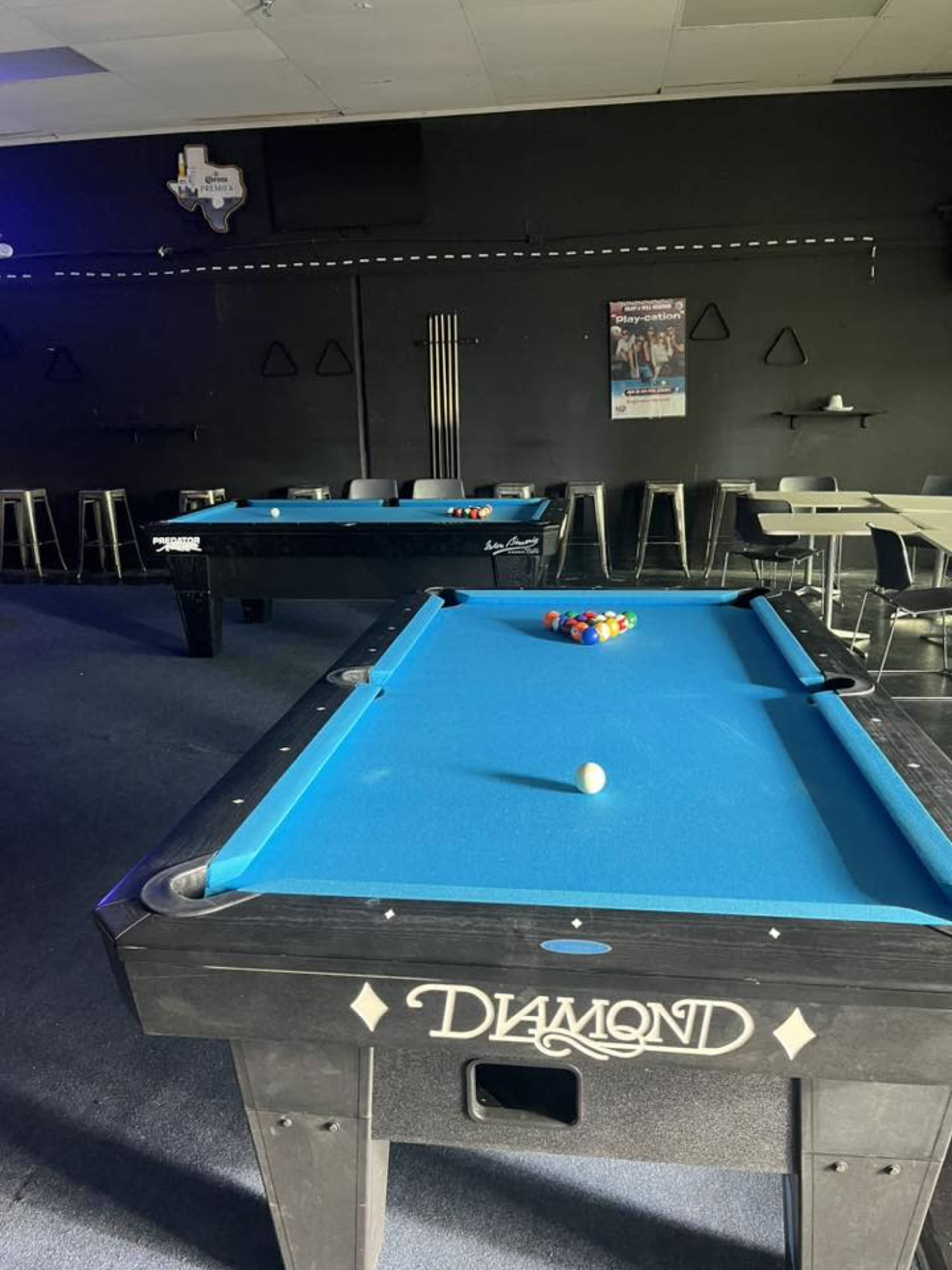 The image shows two pool tables with bright blue felt in a darkened room, with various pool cues and seating in the background.