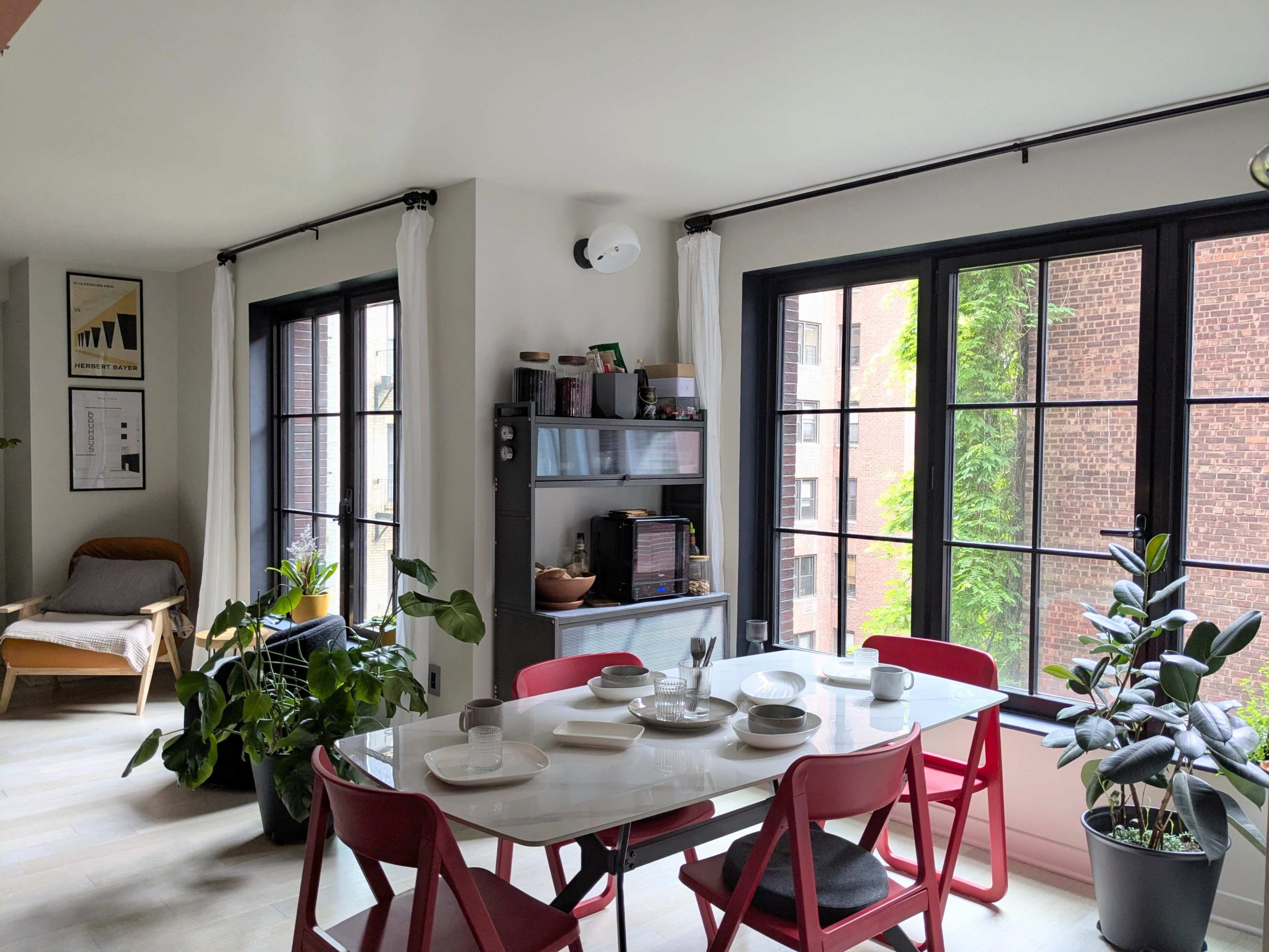 The image shows a dining area with a white table set for a meal, surrounded by red chairs and large windows allowing light to enter, complemented by potted plants.