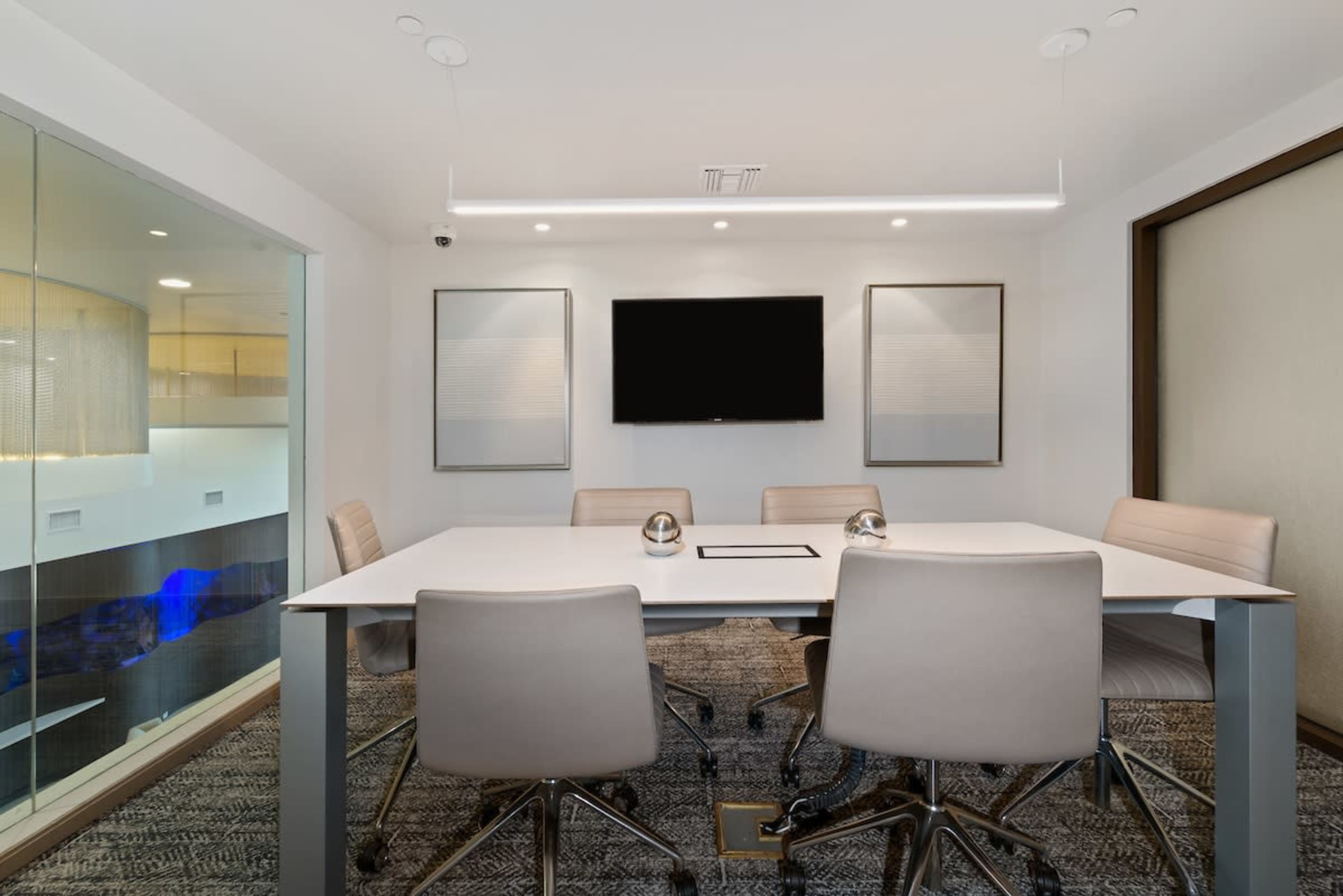 A modern conference room with a white table, four chairs, and a wall-mounted television.