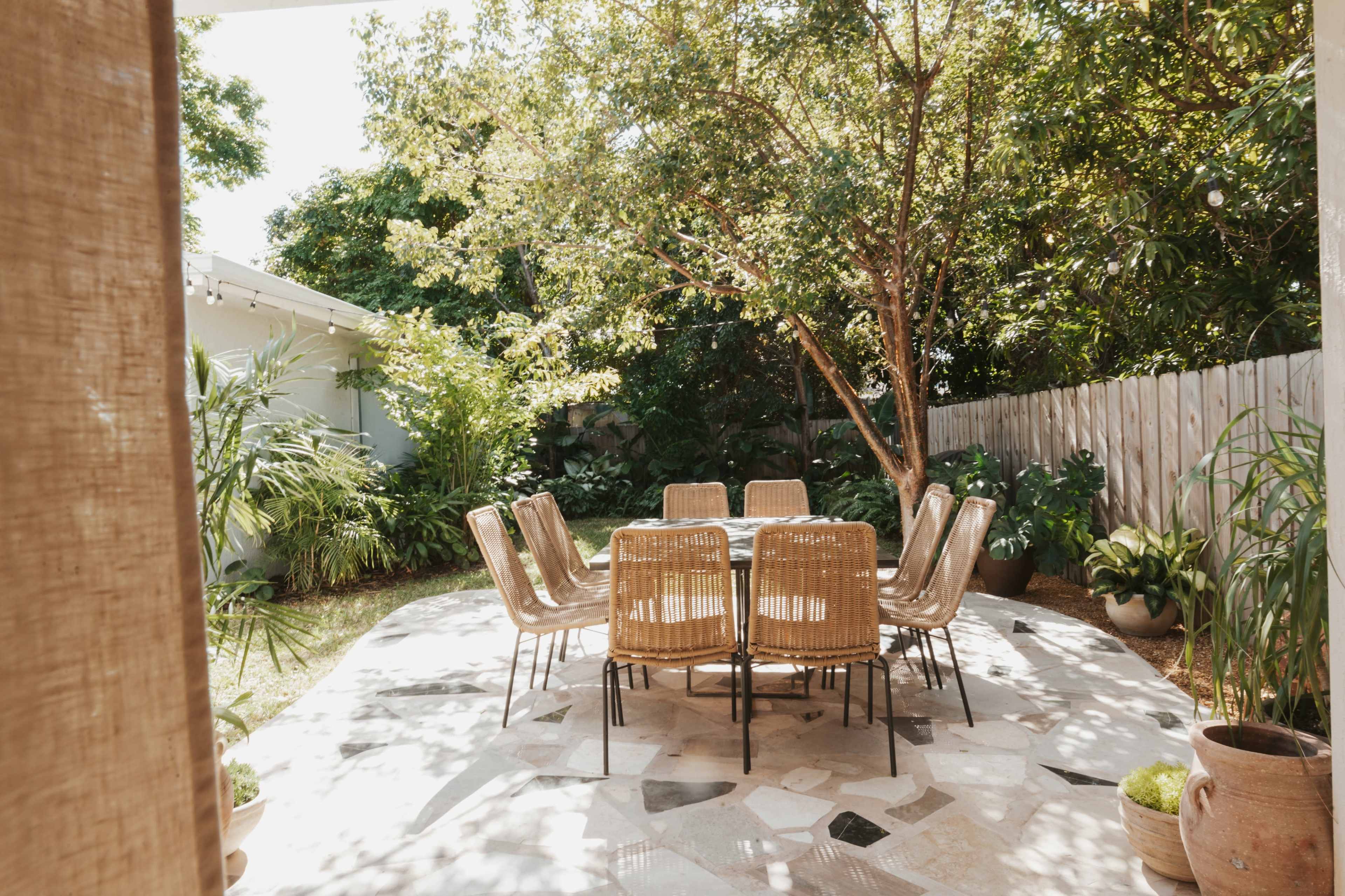 A circular outdoor dining table surrounded by wicker chairs is set in a garden filled with lush greenery and trees.