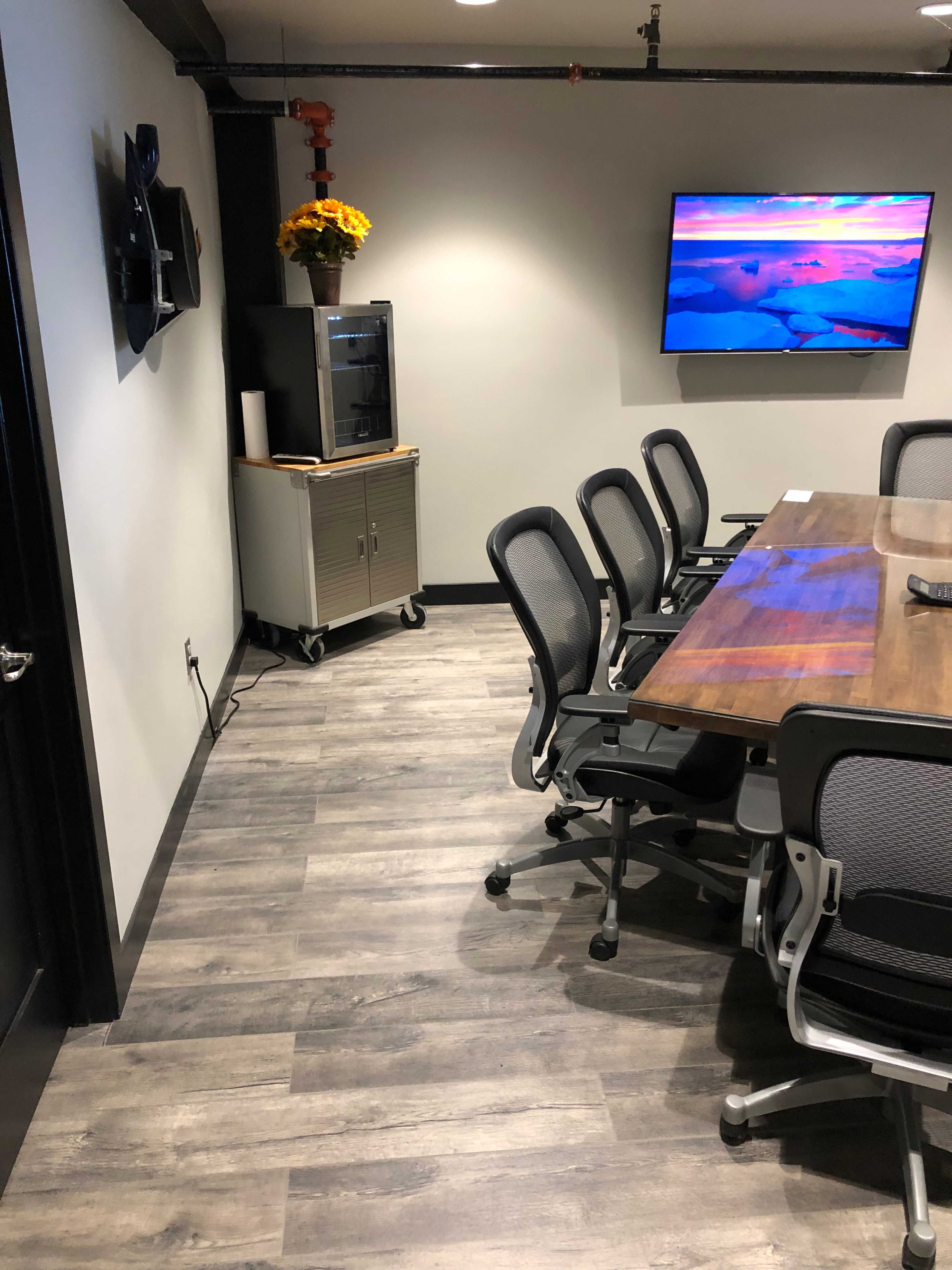A modern conference room with a large wooden table, ergonomic chairs, a television mounted on the wall, and a cabinet with a flower arrangement.