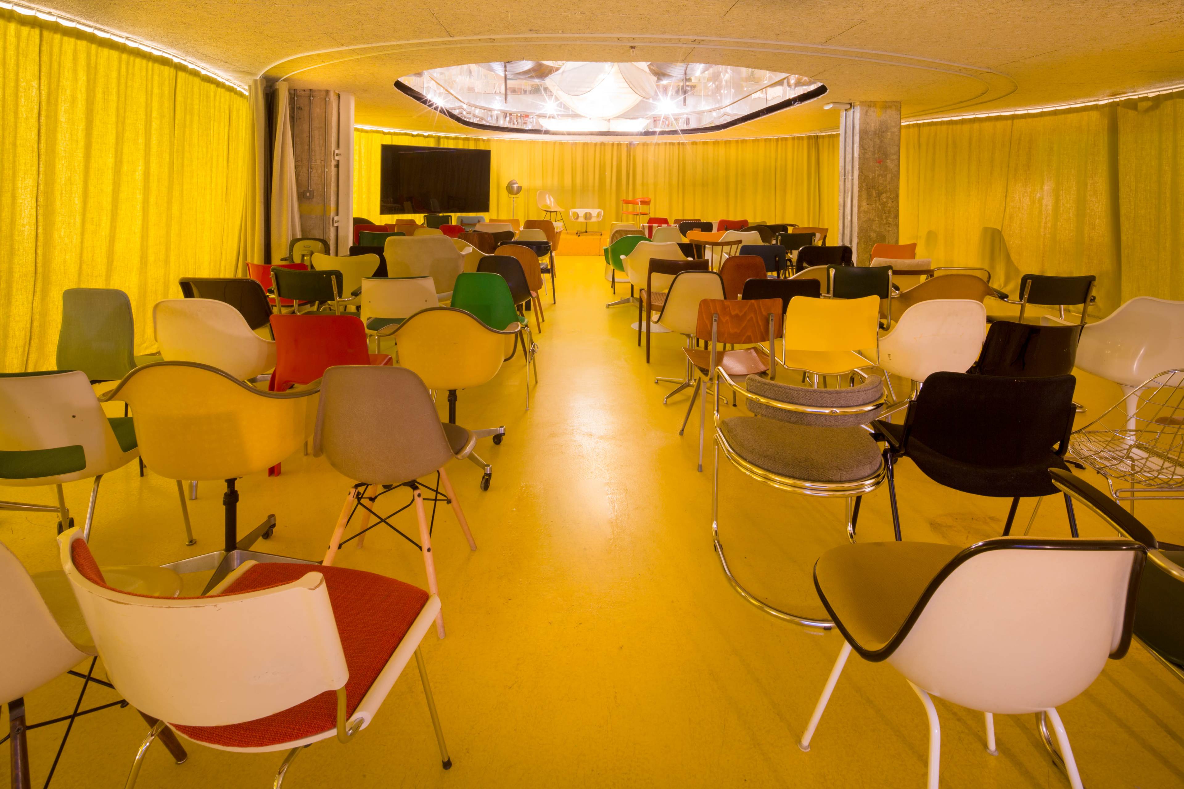 A room filled with an array of chairs in various colors, arranged facing a stage area under a yellow curtain.