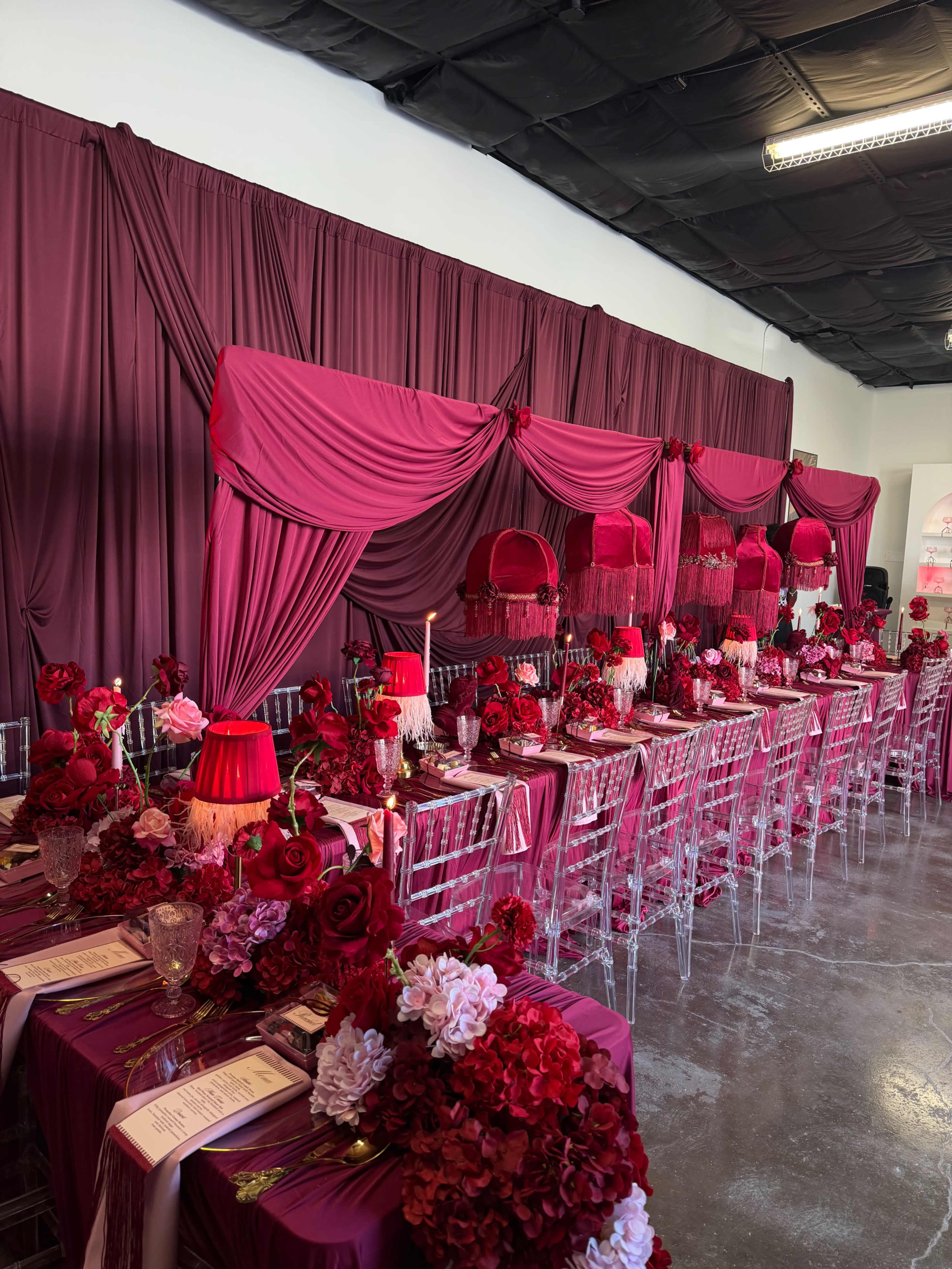 The image shows a lavish dining setup with tables adorned in deep red and pink fabrics, surrounded by decorative floral arrangements and elegant glassware.