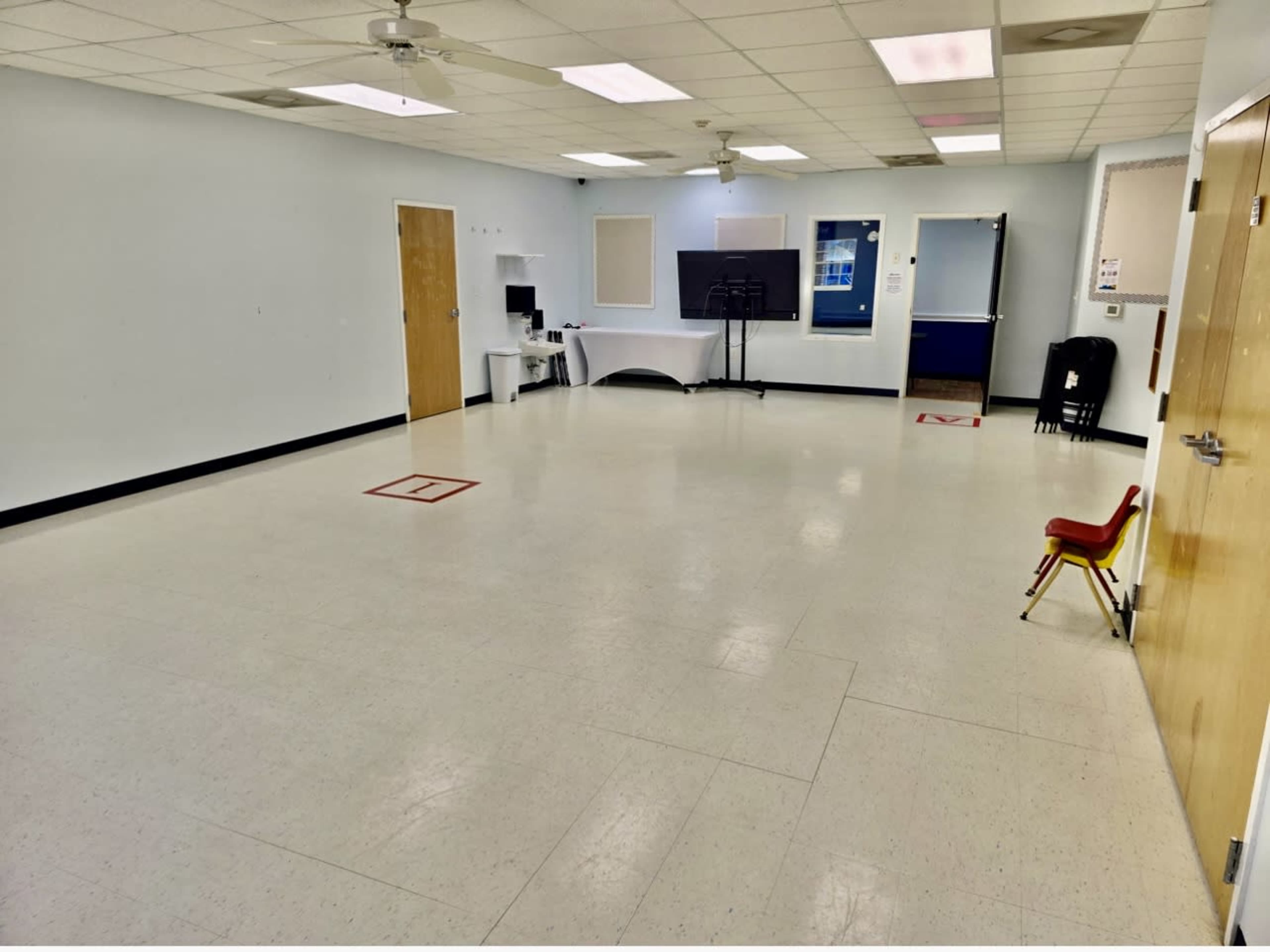 The image shows a large, empty room with light-colored walls, a few doors, a computer setup, and a single red chair placed against one wall.