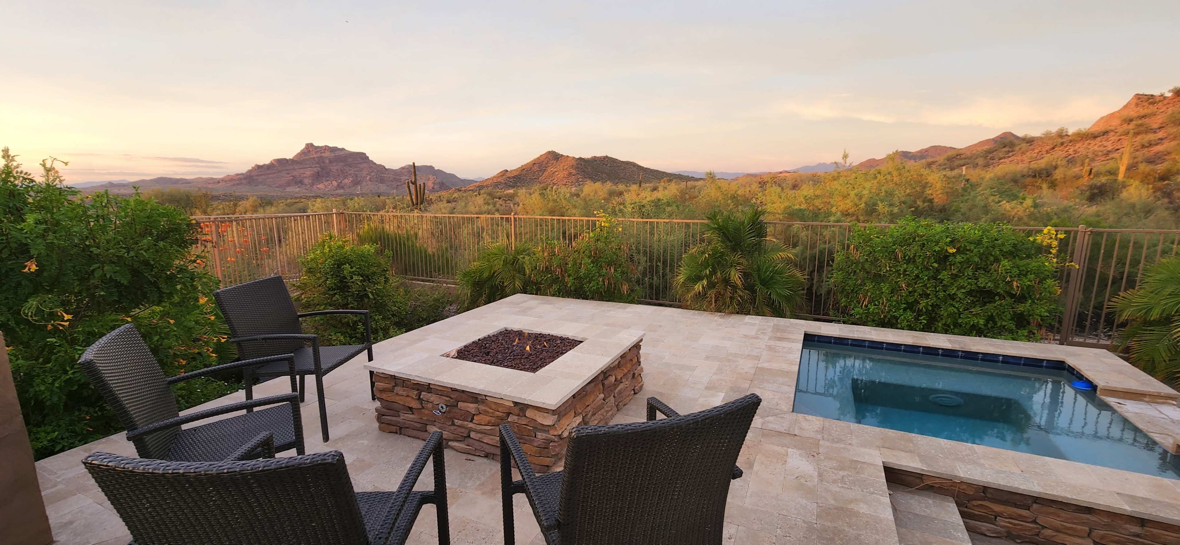 A patio area with four chairs around a stone fire pit and a small jacuzzi, overlooking a desert landscape at sunset.