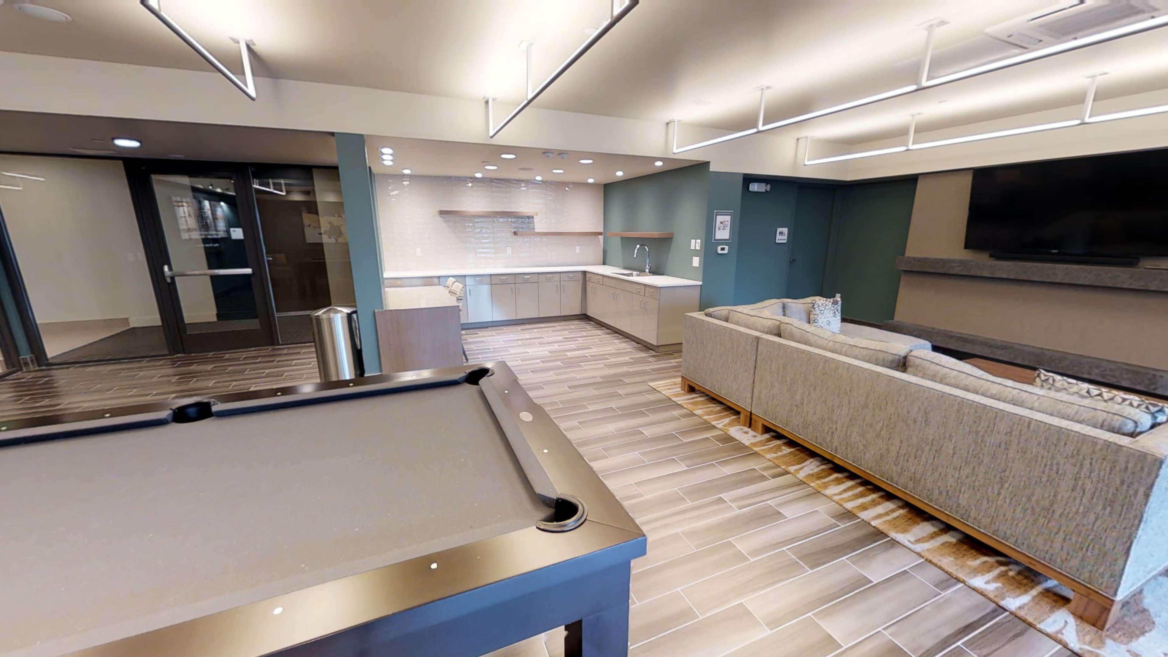 The image depicts a modern lounge area with a pool table in the foreground, a sofa, and a kitchenette in the background.