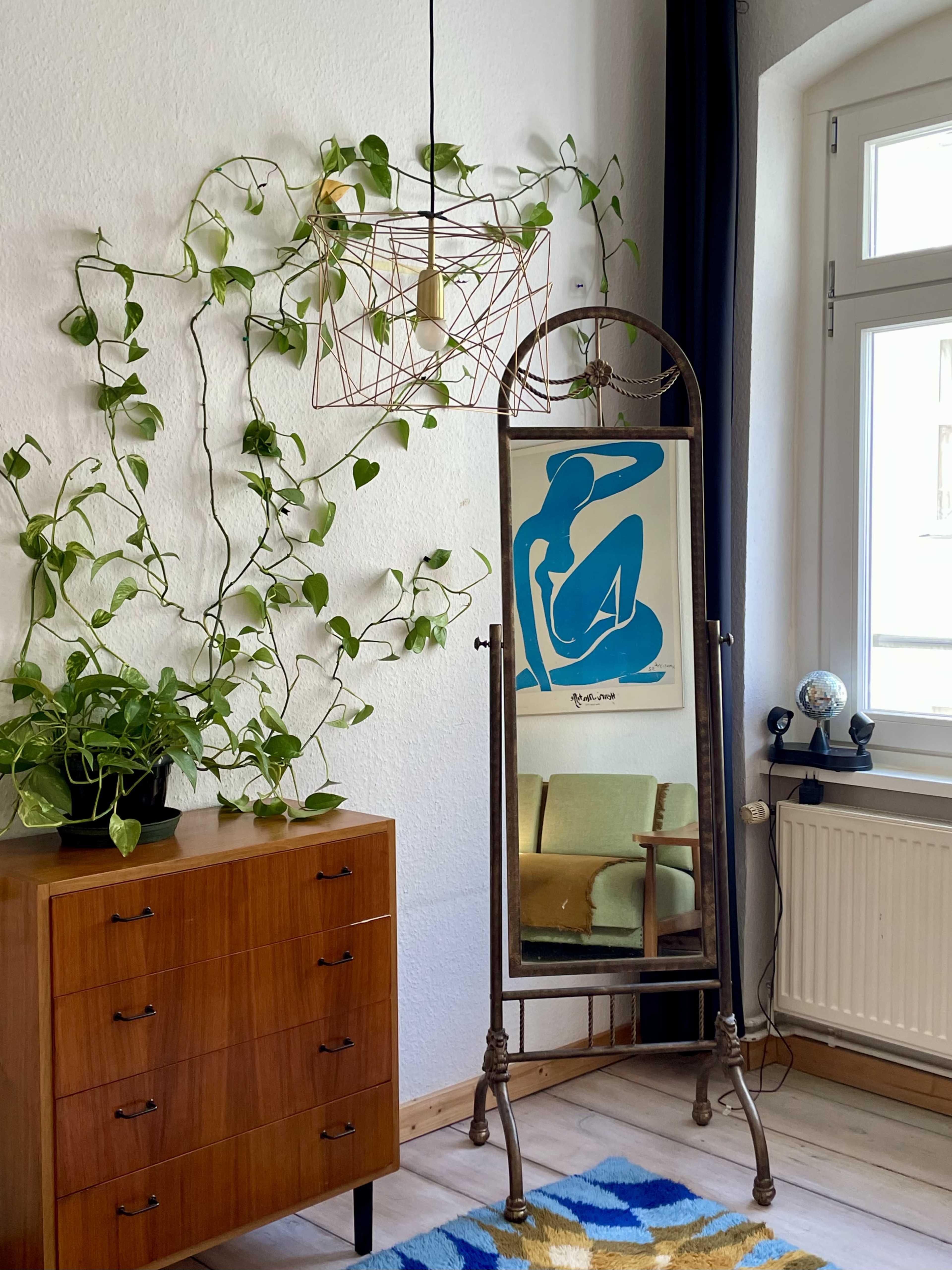 The image shows a corner of a room featuring a full-length mirror, a decorative plant, a pendant light, and a wooden dresser.